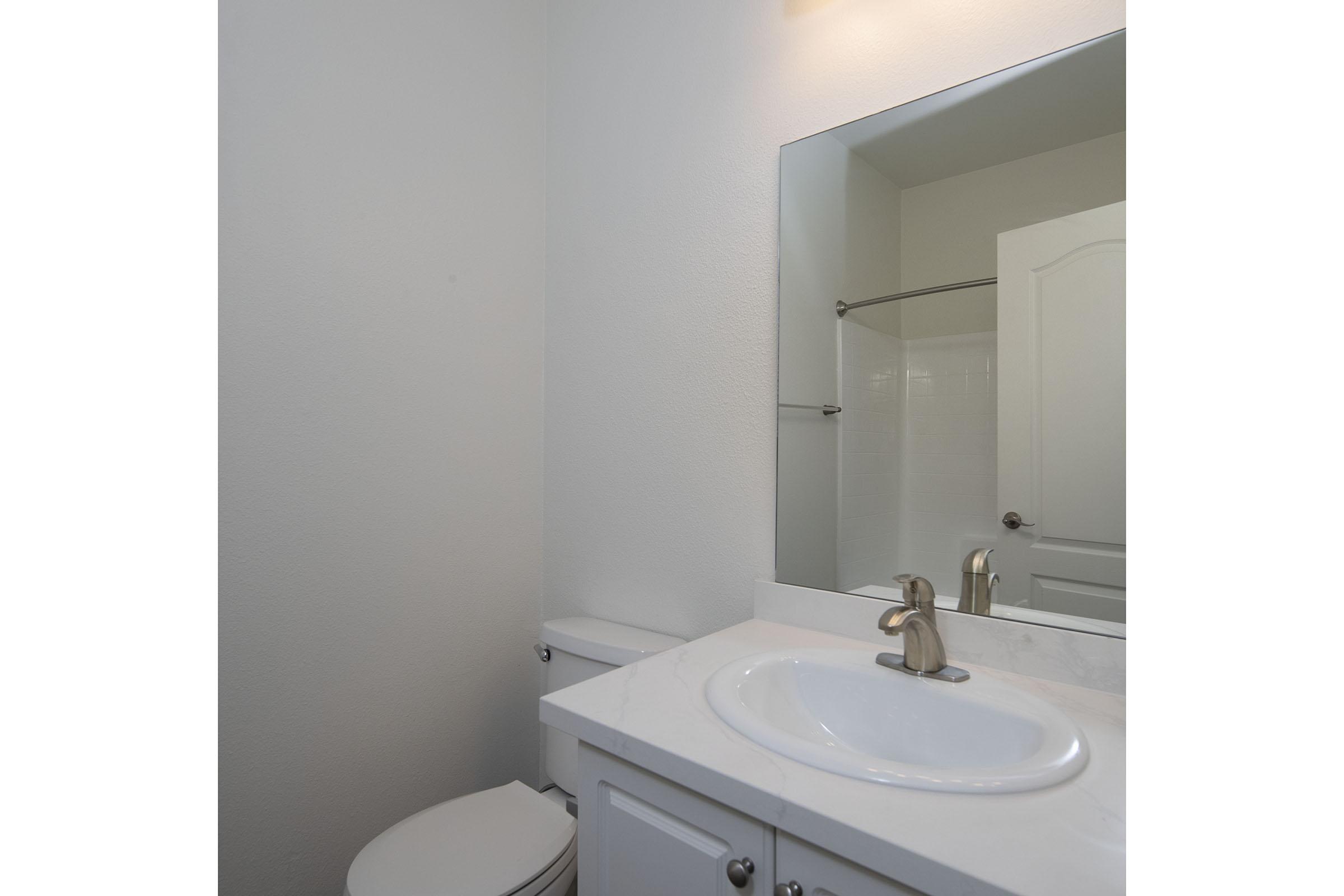 A clean, modern bathroom featuring a white vanity with a round sink, a large mirror above it, and a chrome faucet. The walls are plain and light-colored, and there is a toilet visible to the left. A shower area is in the background, suggesting a simple yet functional design.