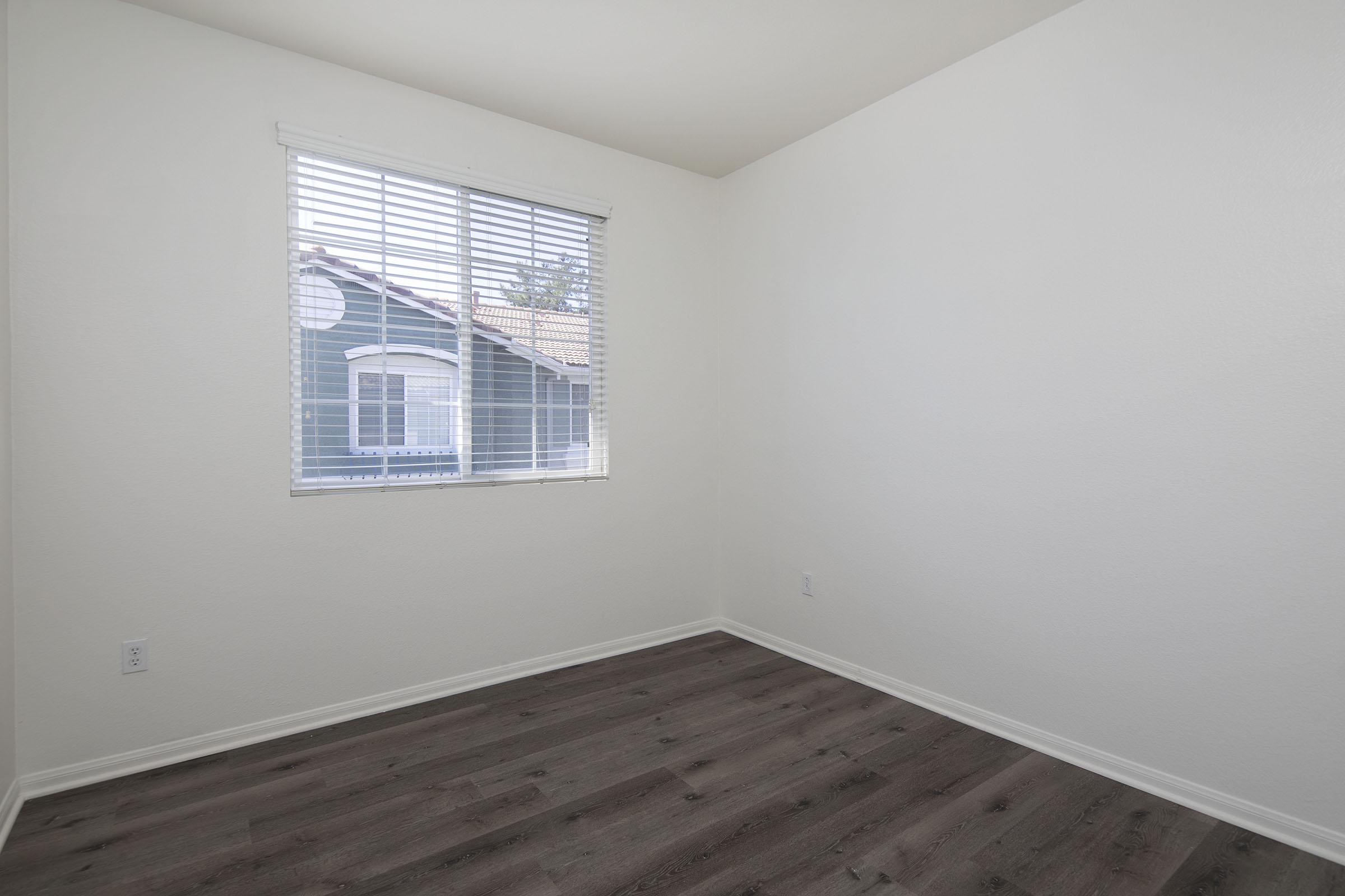 Empty room with light-colored walls, a window with blinds allowing natural light, and a hardwood floor. The window overlooks a house with blue siding outside. The space is uncluttered and ready for furniture.