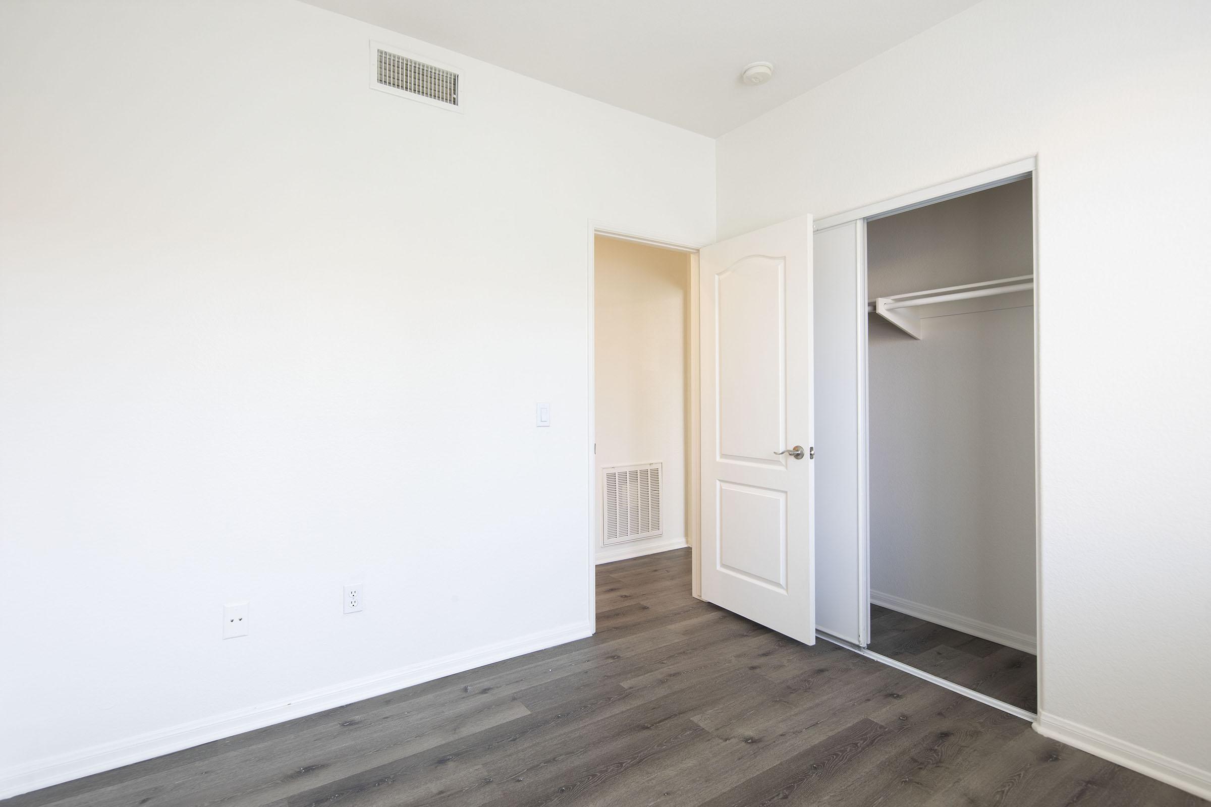 A simple, empty room with light-colored walls and a laminate floor. A closet is visible to the right, featuring sliding doors. The space has a single door leading to another room and a small vent on the wall, creating a bright and airy atmosphere.