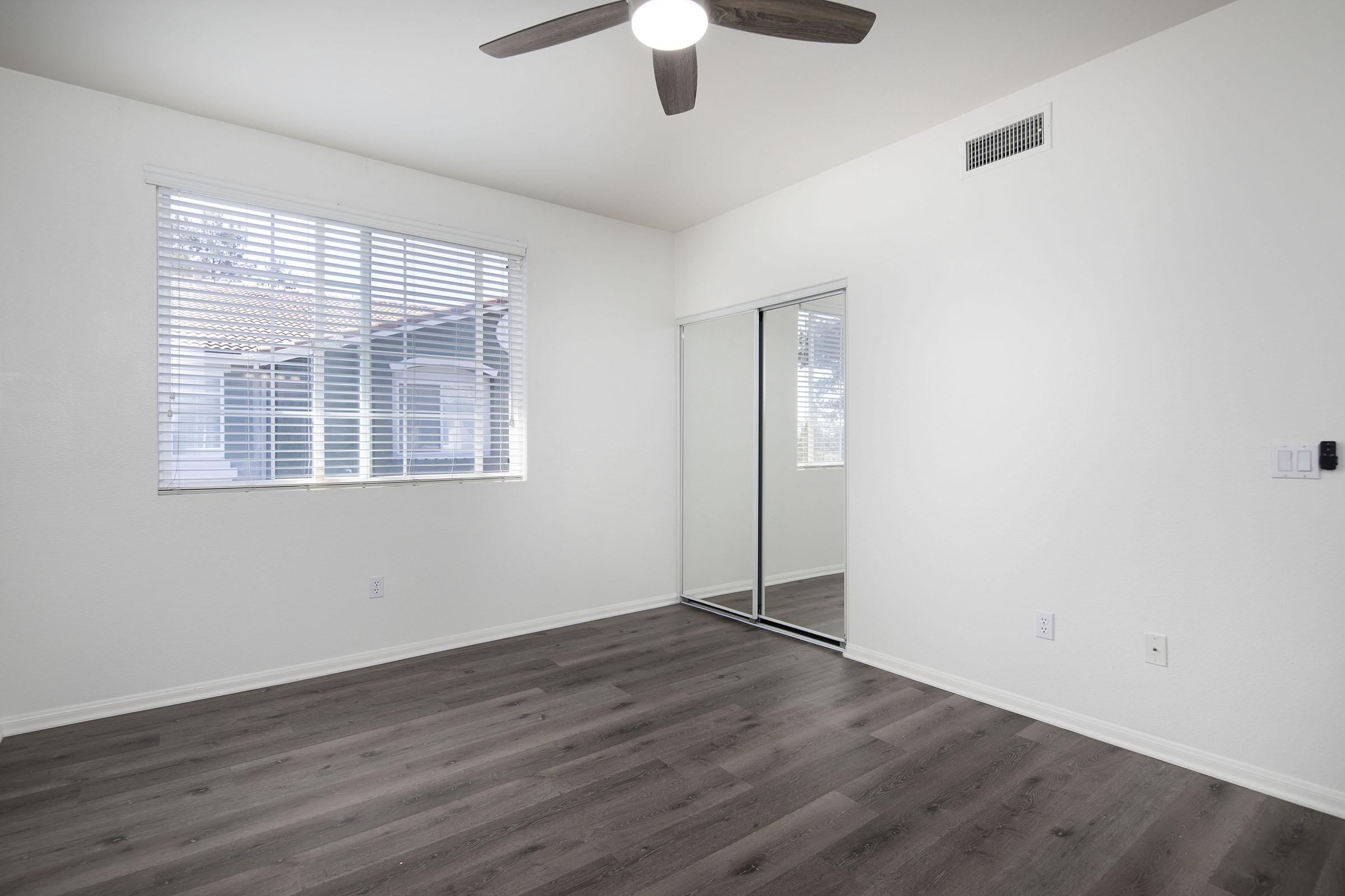 A bright, empty room featuring a ceiling fan, large window with blinds, and a mirrored closet. The walls are painted white, and the floor is covered in light wood laminate. Natural light fills the space, creating a clean and inviting atmosphere.