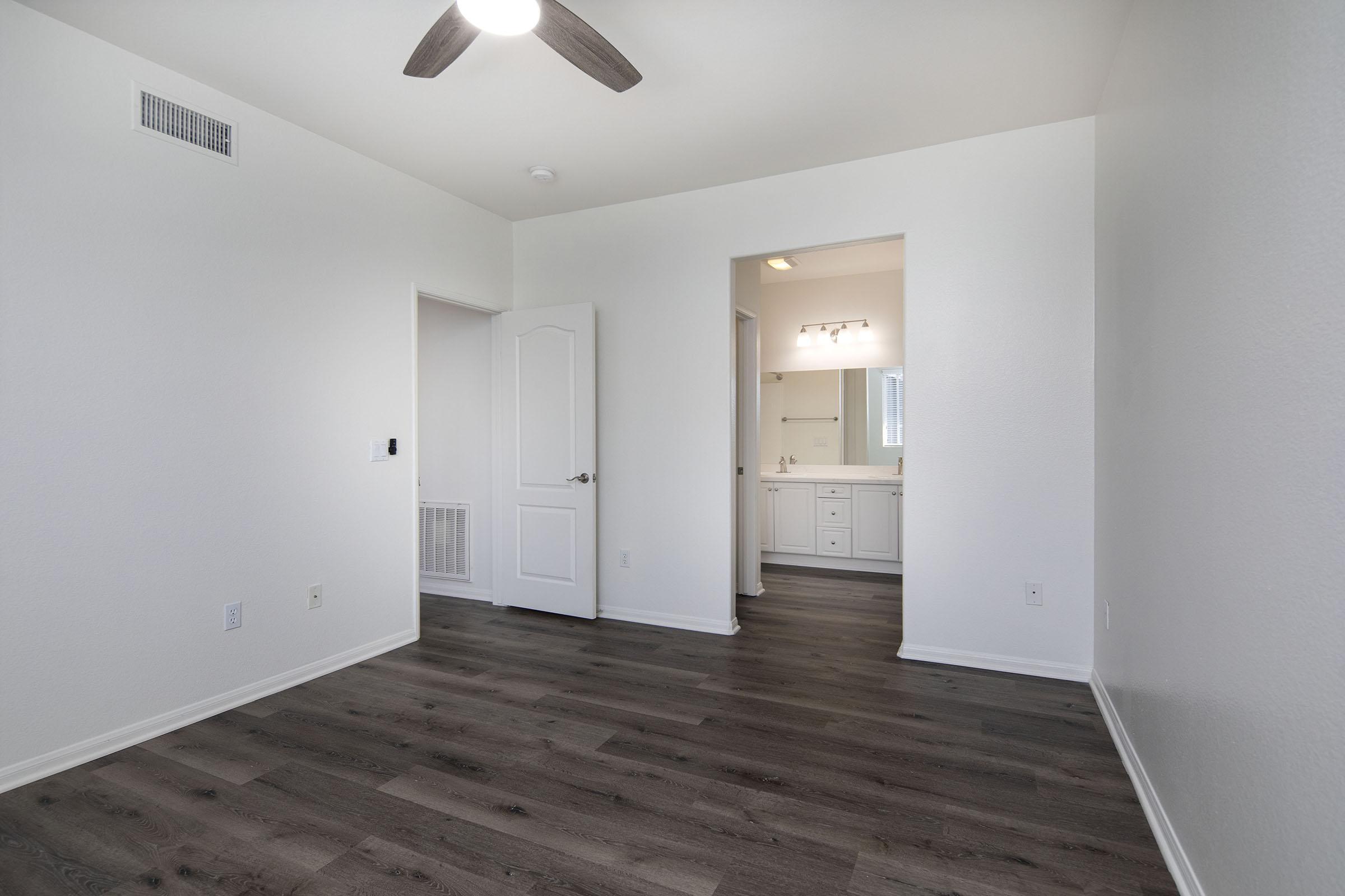 A spacious, empty room with light-colored walls and a modern ceiling fan. The flooring is dark wood, and a doorway leads to a bathroom area featuring a sink and mirror. Natural light enters the space from a window, creating a bright and airy atmosphere.