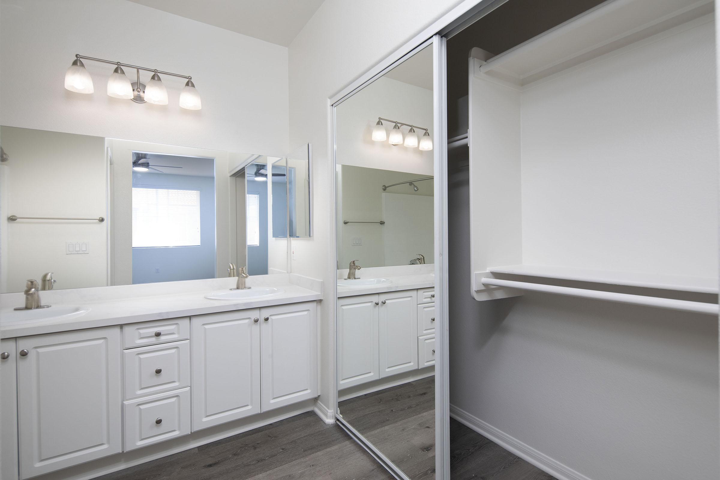 A modern bathroom featuring a double sink vanity with white cabinetry, large mirrors, and a bright light fixture. The space includes a walk-in closet with shelving and a neutral color scheme, complemented by natural light from a nearby window. The flooring is designed with a stylish, contemporary look.