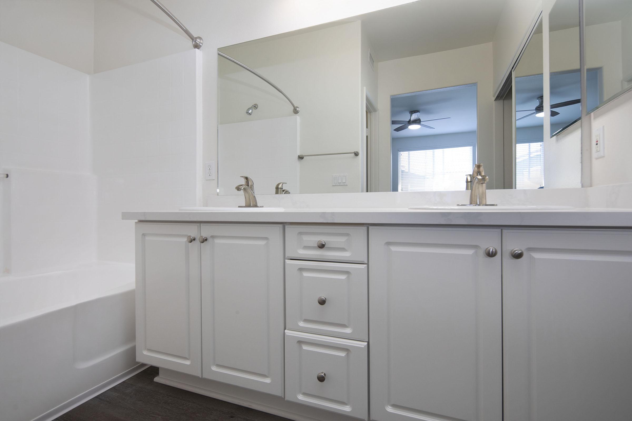 A modern bathroom featuring a white double vanity with chrome faucets, light-colored cabinetry, and a large mirror. The space includes a bathtub with a white surround, a shower rod, and a window allowing natural light, enhancing the clean and bright atmosphere.