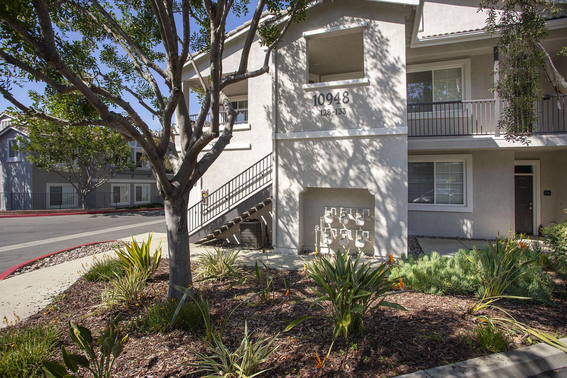 A multi-story residential building with a light gray exterior and landscaping featuring various plants and shrubs. The building has a set of stairs leading to the second floor, and an address plaque reading "10948" near the entrance. Sunlight casts shadows on the walkway and surrounding area.