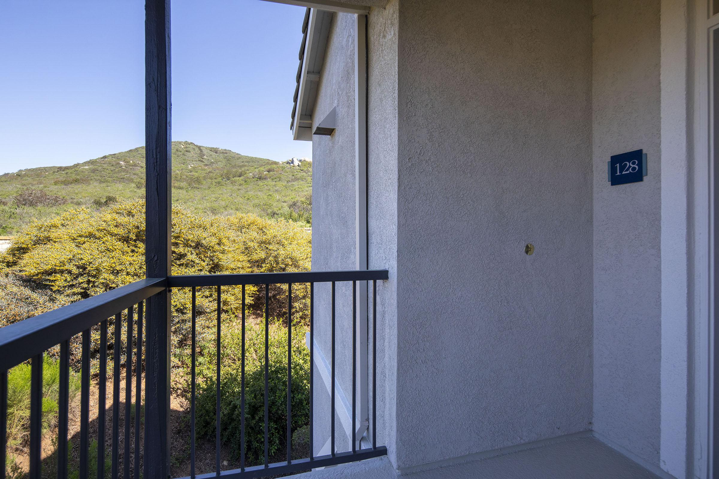 View from a balcony featuring a painted wall with the number 128, overlooking a lush green hillside under a clear blue sky. The scene captures a peaceful outdoor space surrounded by nature.