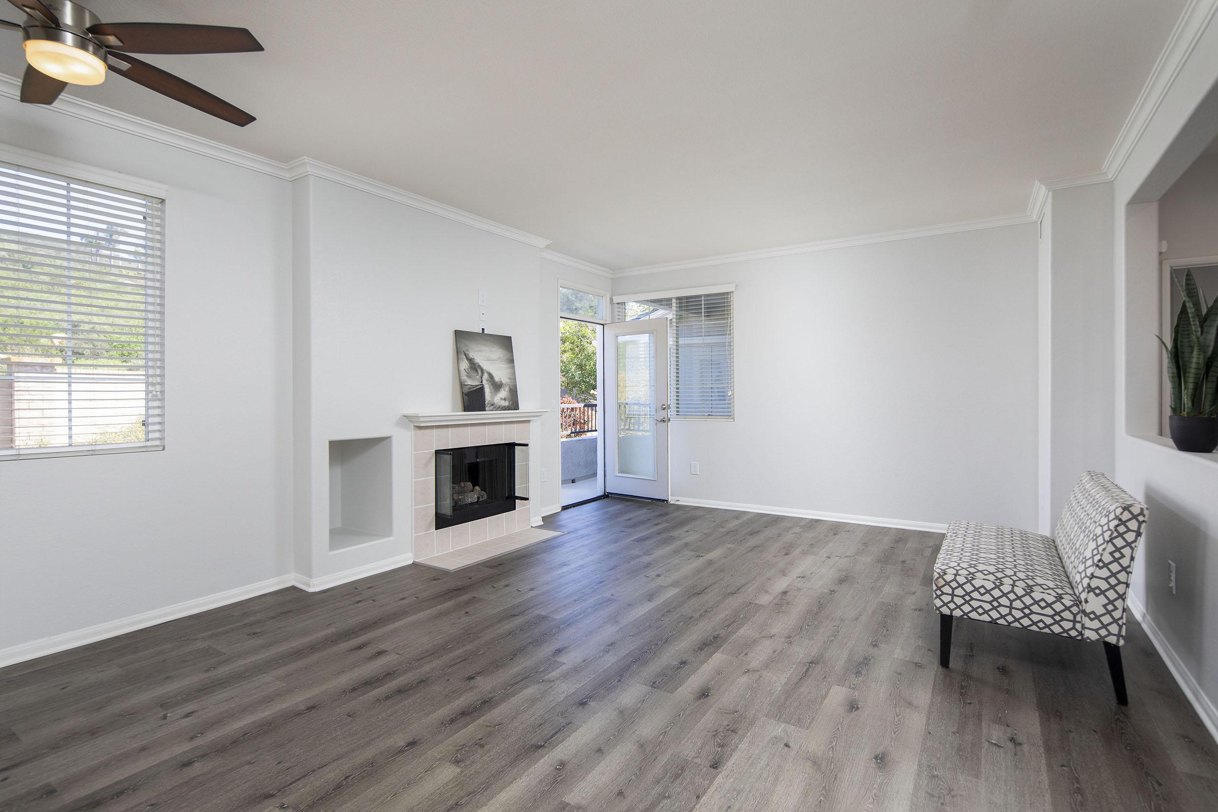 A bright and airy living room featuring light-colored walls, a modern ceiling fan, and large windows. A decorative fireplace is situated on one wall, and a stylish, patterned bench is placed against another wall. The flooring is a sleek, gray wood laminate, creating a contemporary and inviting atmosphere.