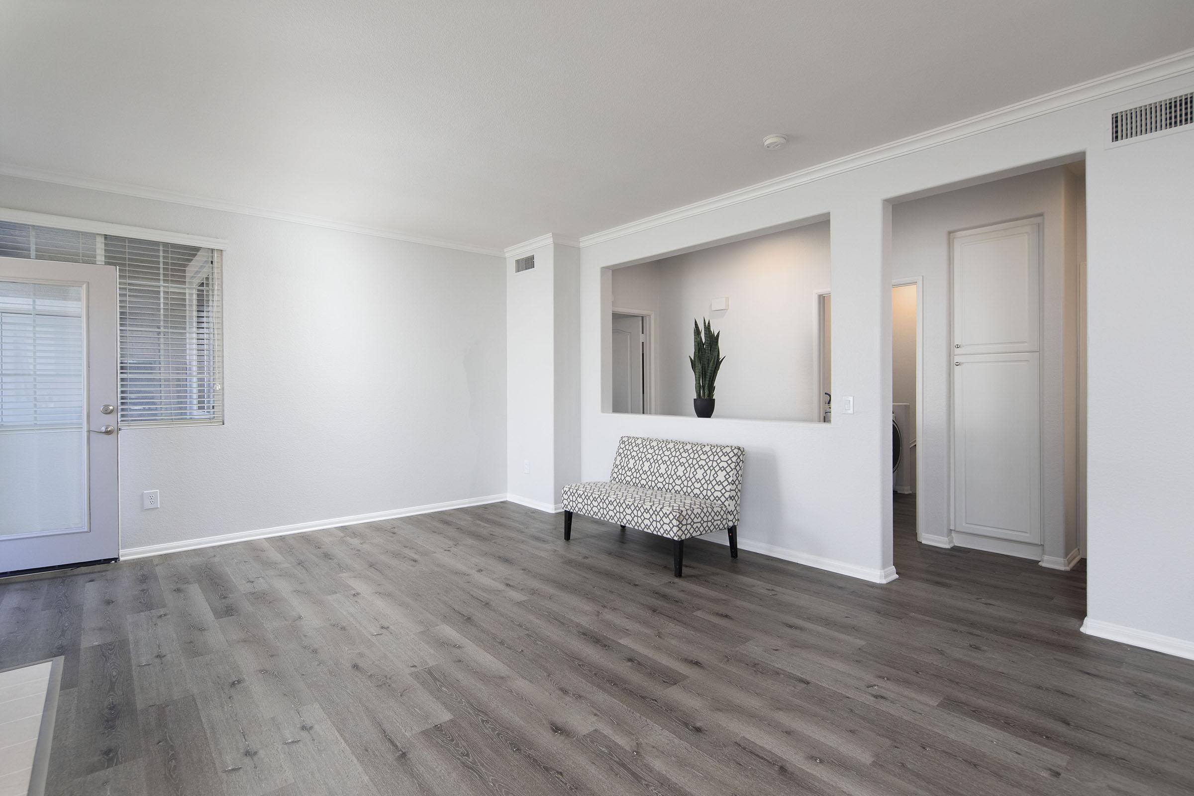 A spacious, modern living room with light gray walls and wood-like flooring. A small, patterned bench is placed against one wall. To the left, there is a door leading outside, while a doorway in the back opens to another room. A potted plant is positioned on the bench, adding a touch of greenery.