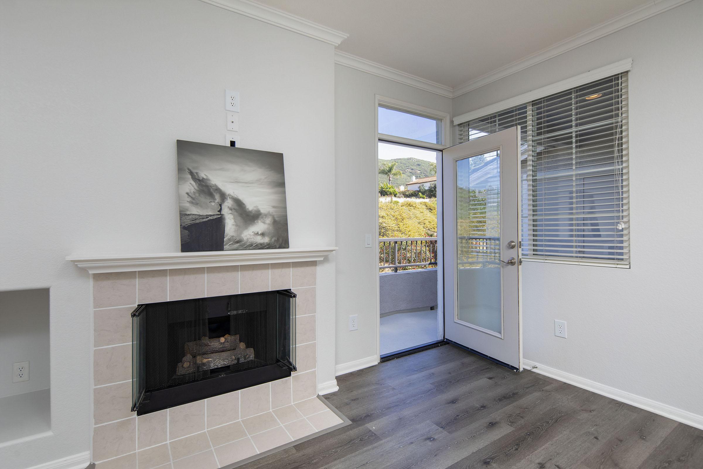 A bright, modern living space featuring a cozy fireplace with a glass front, a large window letting in natural light, and sleek, wood-style flooring. A black-and-white artwork hangs above the fireplace. The door in the corner opens to a balcony, offering views of green foliage and hills outside.