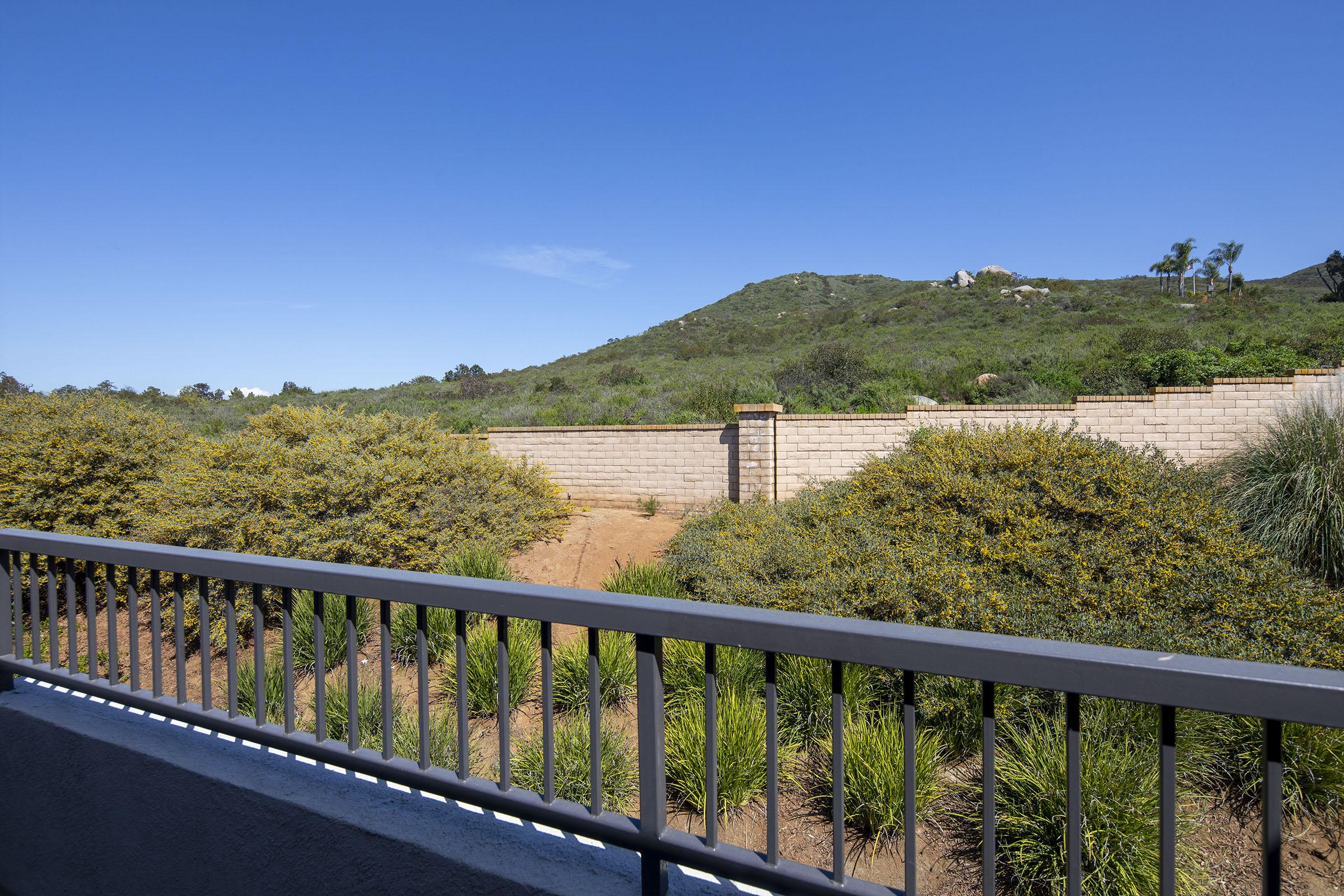 A view from a balcony showcasing a landscape with a low stone wall and lush greenery. In the background, rolling hills are visible against a clear blue sky. The scene conveys a peaceful and natural setting, ideal for relaxation.