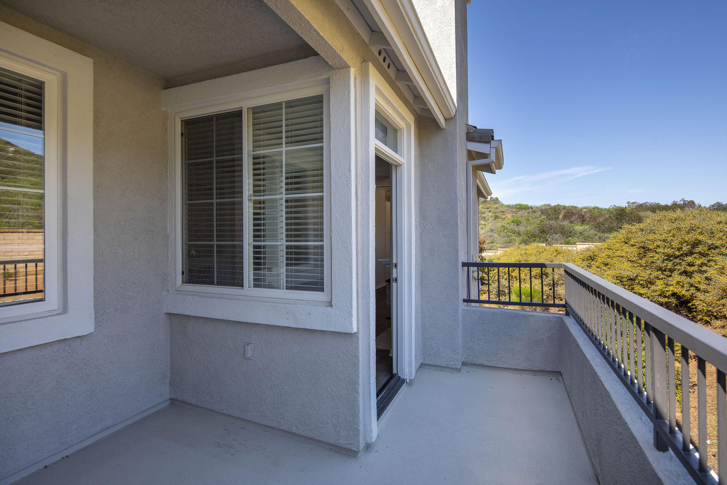 A sunny balcony with a view, featuring a large window with white blinds and a railing. The surrounding area shows greenery and hills in the background under a clear blue sky. The floor is light-colored concrete, creating a spacious and inviting outdoor space.