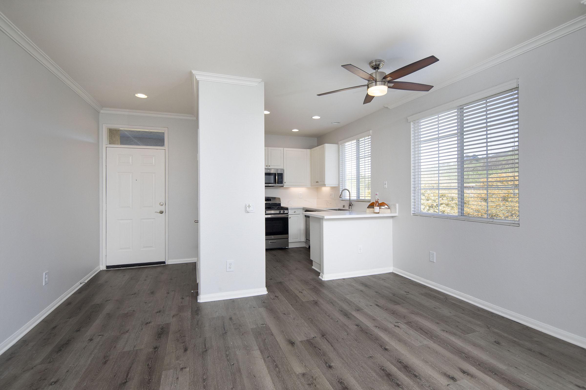 A modern, empty living area featuring light walls and a spacious layout. On the left, a front door leads outside. To the right, a contemporary kitchen with stainless steel appliances and a small island is visible. Large windows allow natural light, and wooden flooring runs throughout the space.