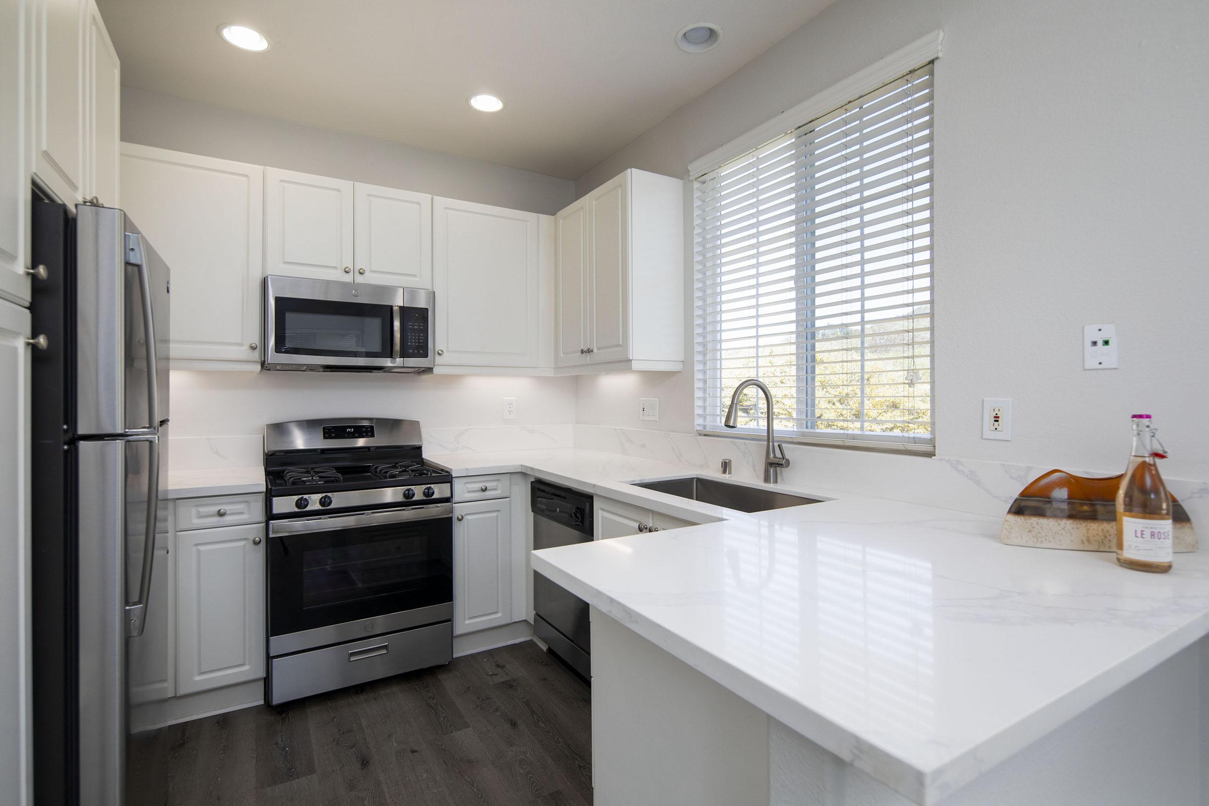 Modern kitchen with white cabinetry, stainless steel appliances including a refrigerator and microwave, a gas stove, and a dishwasher. The countertops are light-colored with a marble finish. A window with blinds allows natural light to enter, and a bottle of wine sits on the counter.