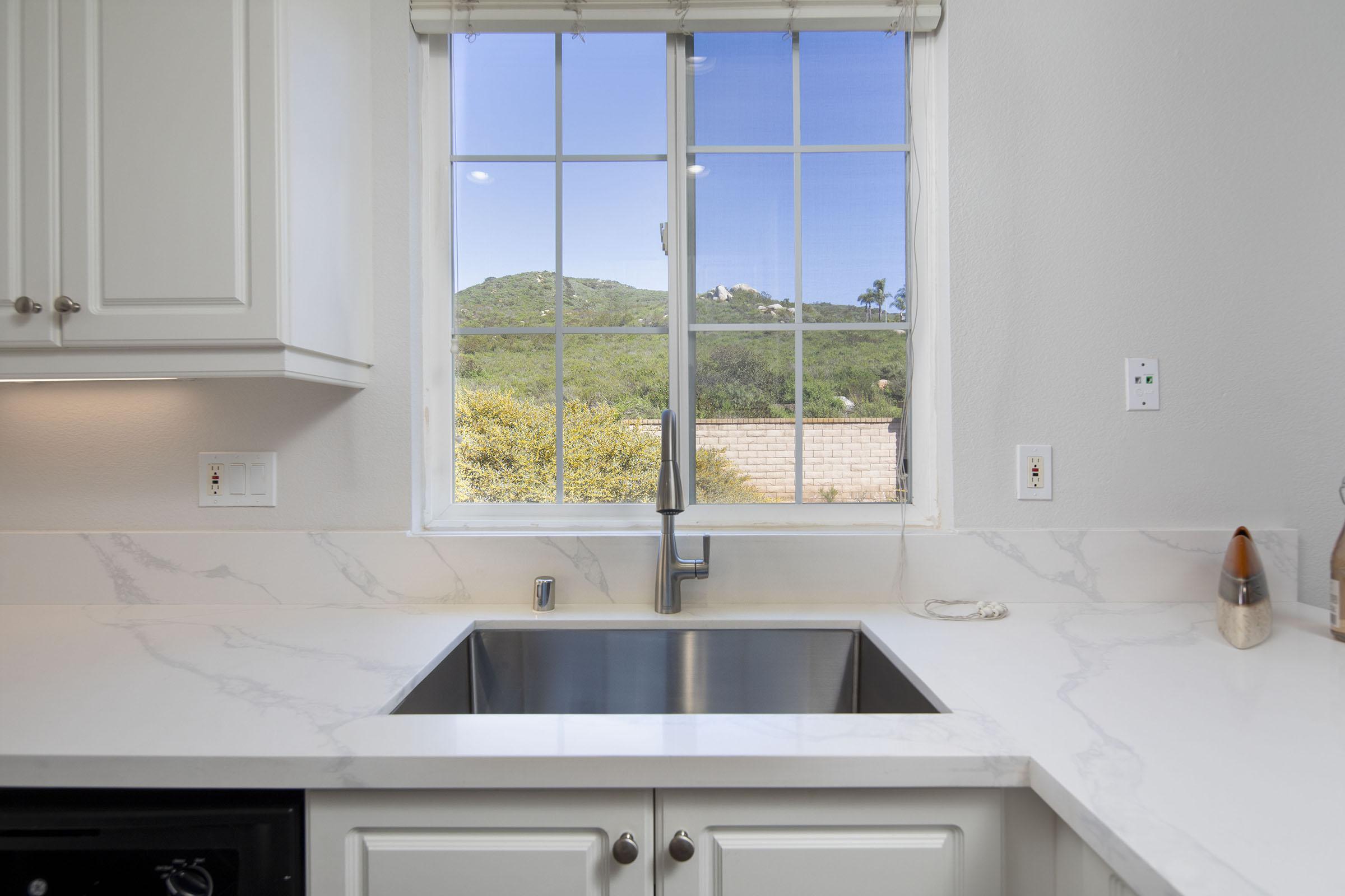 A modern kitchen with a stainless steel sink and marble countertop. In the background, a large window offers a view of green hills and trees under a clear blue sky. The cabinetry is light-colored, adding to the bright and airy feel of the space.