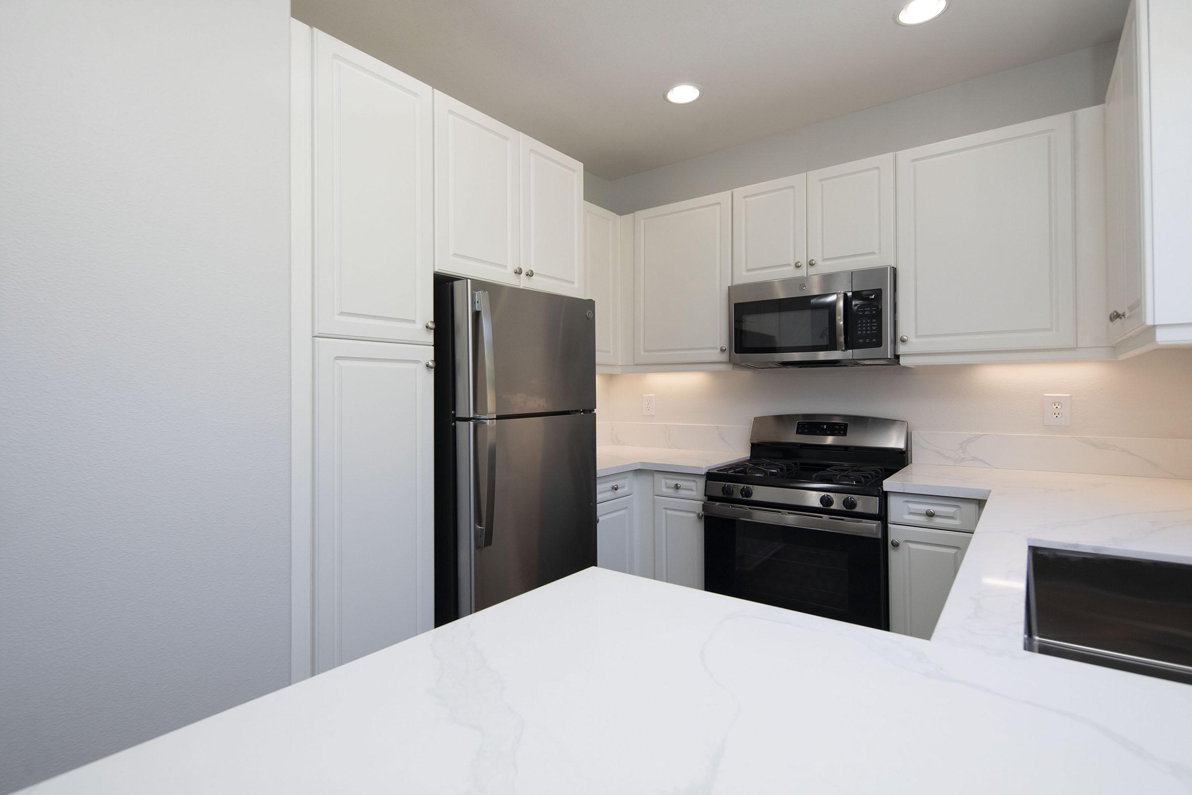 Modern kitchen featuring white cabinetry, stainless steel refrigerator, and microwave. A gas stove is integrated into the countertop, which has a marble-like finish. The space is well-lit with overhead lighting and has a clean, contemporary design.