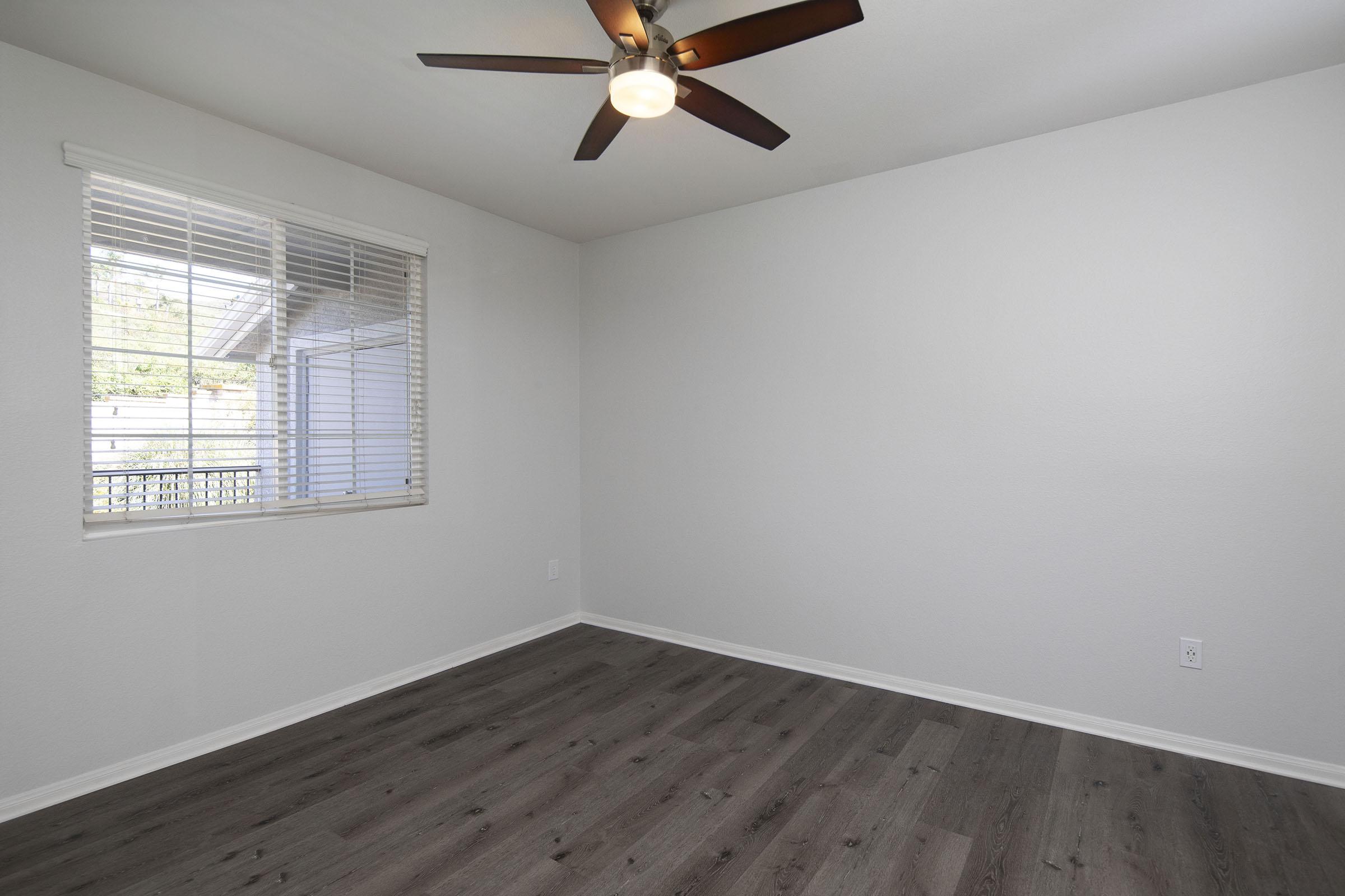 A spacious, empty room with light gray walls and a ceiling fan. It features a window with closed blinds, allowing natural light to brighten the space. The floor is covered in dark laminate, contributing to a modern, clean aesthetic. No furniture is present, emphasizing the room's openness.