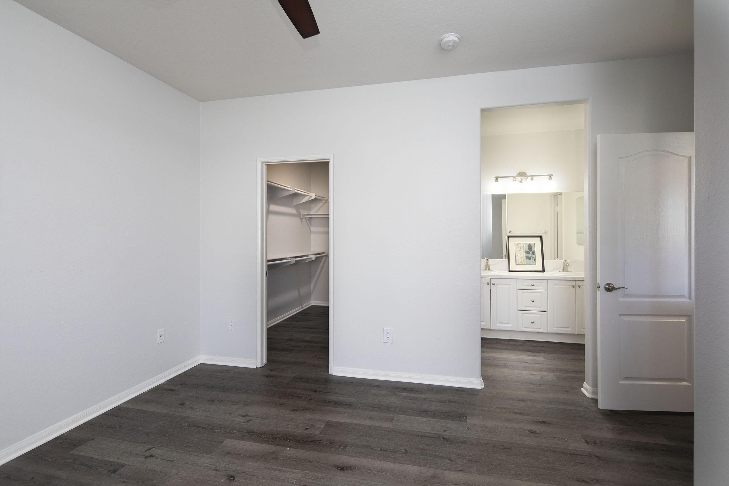 A bright, empty bedroom featuring light-colored walls and wood-like laminate flooring. There's a spacious closet on the left with open shelves, and a doorway on the right leading to a bathroom area with a double vanity and mirror. A ceiling fan is visible, providing a modern touch.