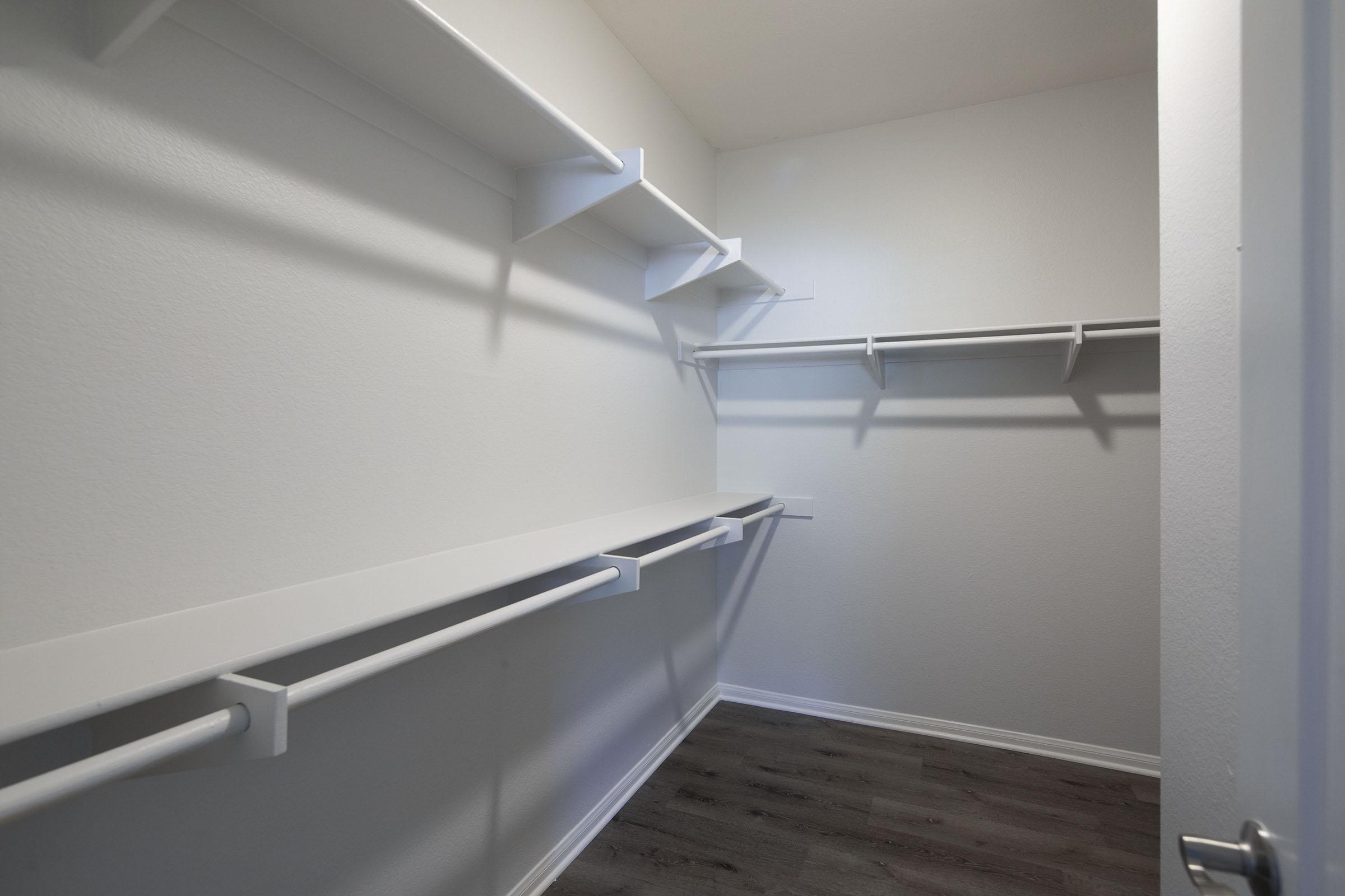 Empty closet with white shelving and hanging rods against light-colored walls. The floor features a dark wood finish, and the overall space appears tidy and well-organized, ready for storage or personal items.