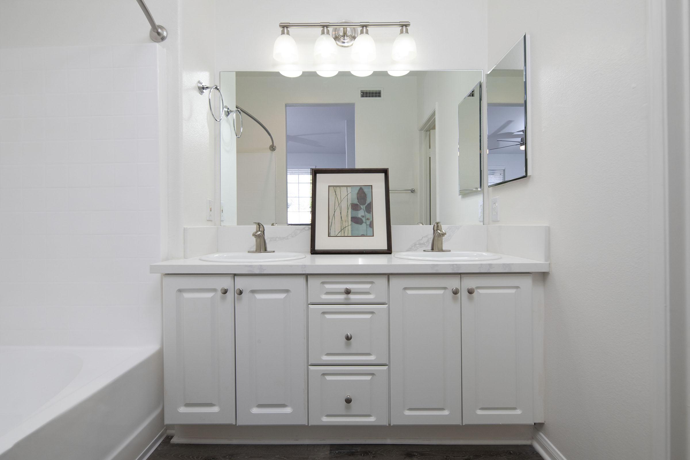 A clean and modern bathroom featuring a dual sink vanity with white cabinets, silver faucets, and a decorative framed piece of art. In the background, there is a bathtub on the left and a large mirror above the sinks, reflecting neutral-colored walls and natural light from a window.