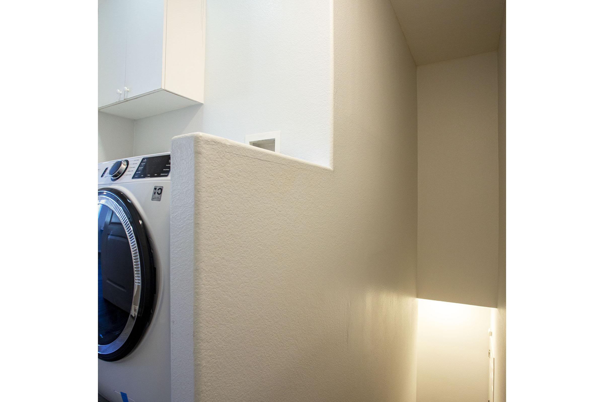 A view of a small laundry area featuring a modern washing machine on the left. The wall is partially covered with a light-colored texture, and there is a small doorway on the right emitting soft light, indicating potential access to another space. The overall ambiance is clean and minimalistic.