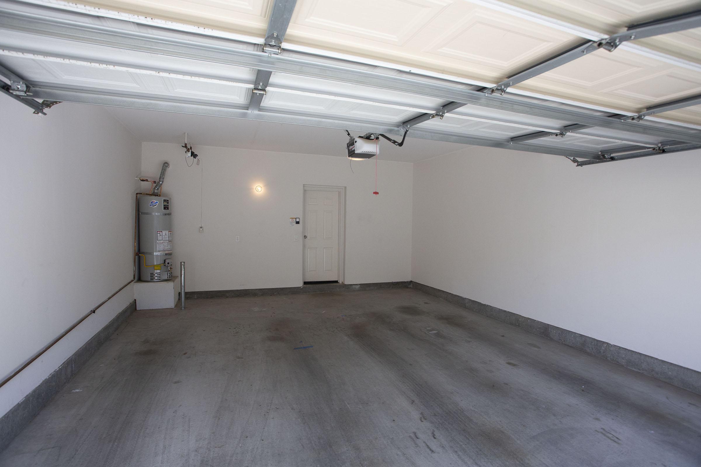 Empty garage with concrete flooring, white walls, and a closed overhead door. A hot water heater is positioned against the left wall, and a single door is visible on the right side. No vehicles or storage items are present in the space.