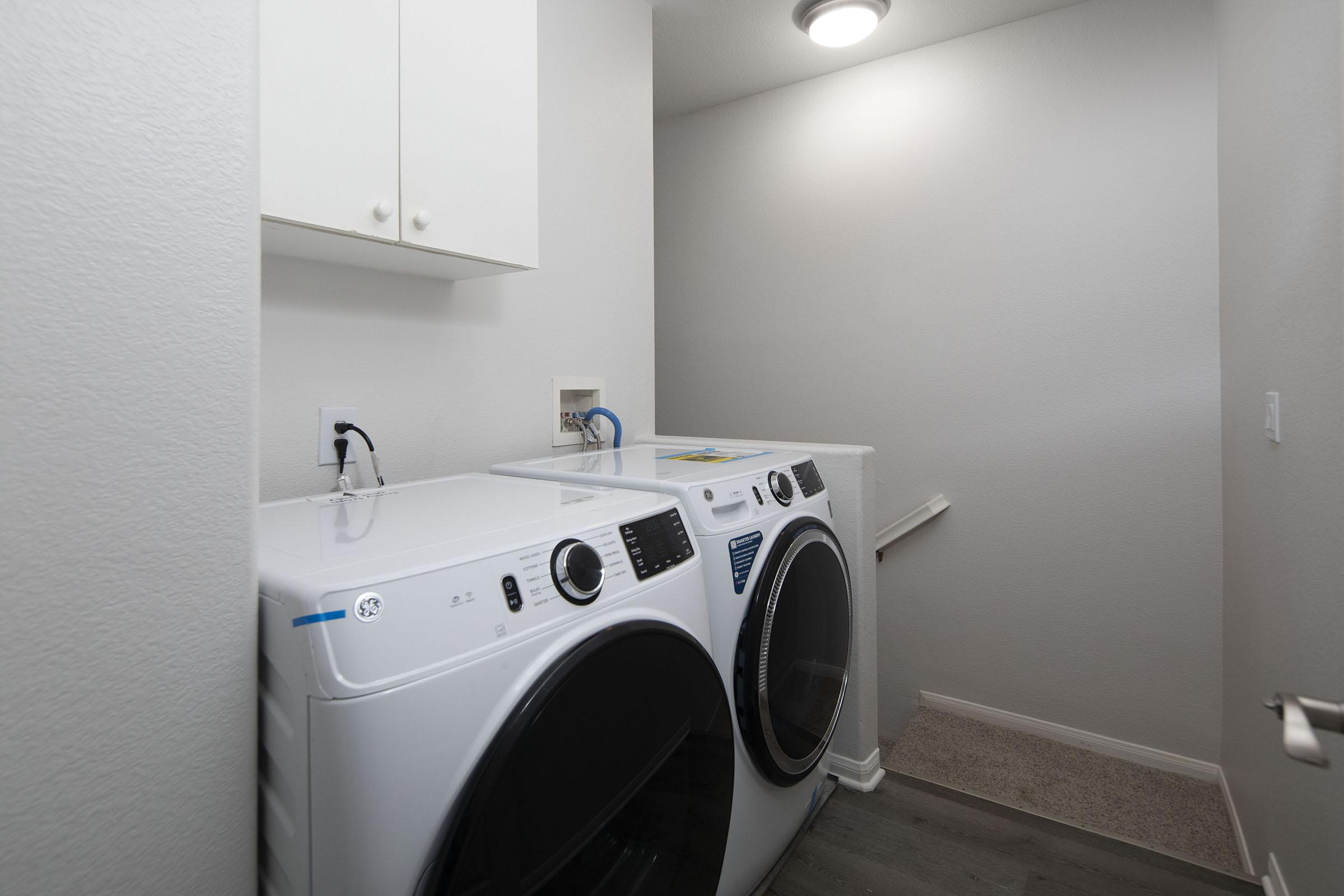 A clean and modern laundry room featuring a pair of stacked white washing machines and dryers. The appliances are positioned against a light grey wall, with a small shelf above them. The floor is carpeted, and there is a light fixture on the ceiling, creating a bright and airy atmosphere.