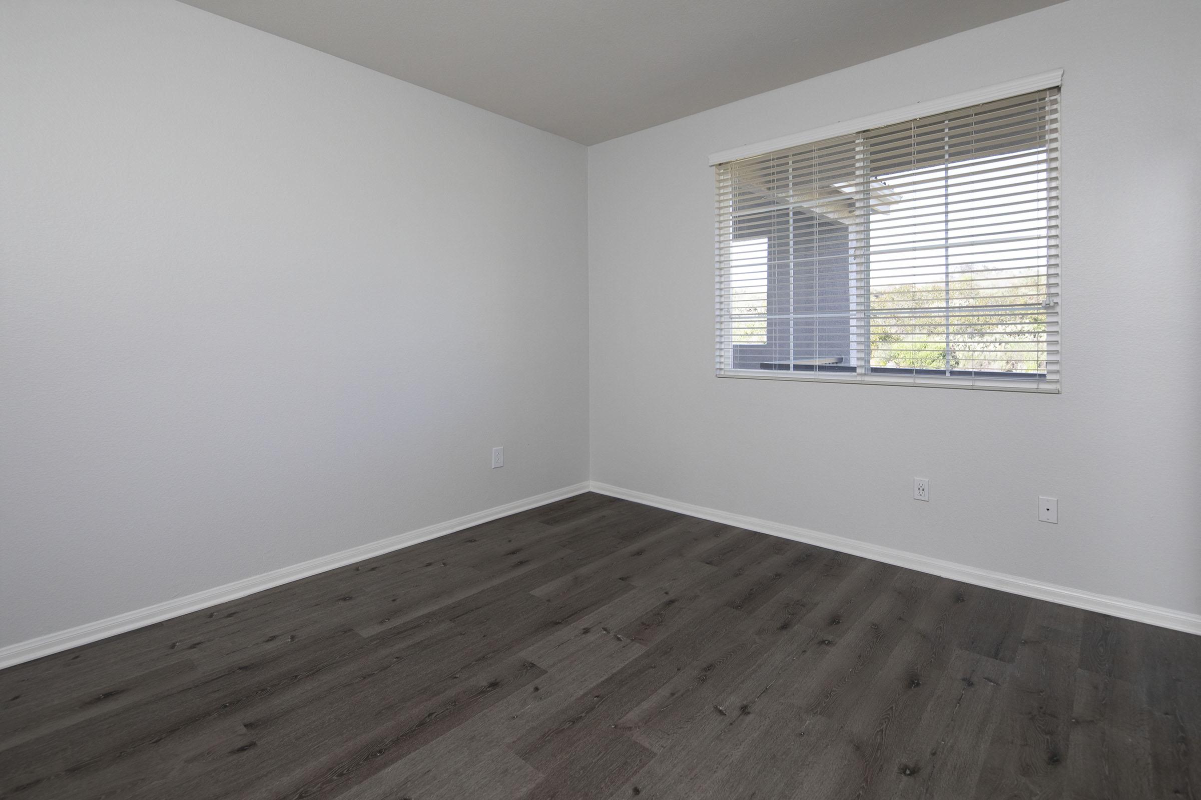 A spacious, empty room with light gray walls and a large window featuring white blinds. The floor is made of dark laminate wood, and there are no furniture pieces in sight, creating a clean and minimalist atmosphere. Natural light enters through the window, illuminating the space.