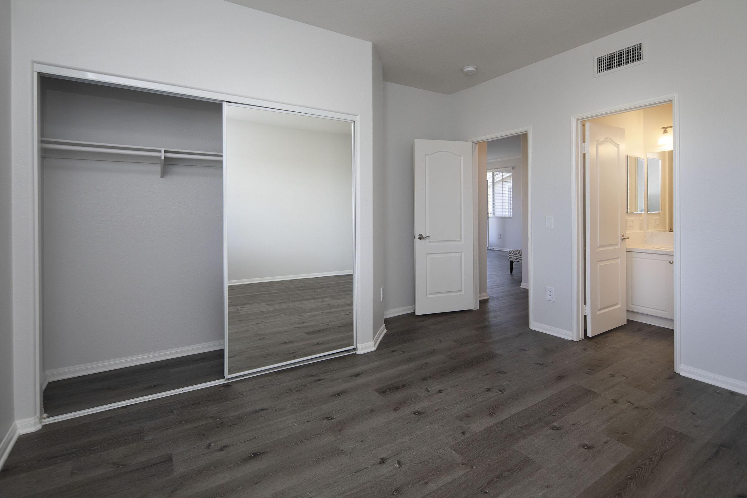 A well-lit interior view of a room featuring a mirrored closet door. The flooring is a dark wood finish, and there is a doorway leading to a bathroom on the right. The walls are painted light gray, creating a spacious and modern atmosphere.