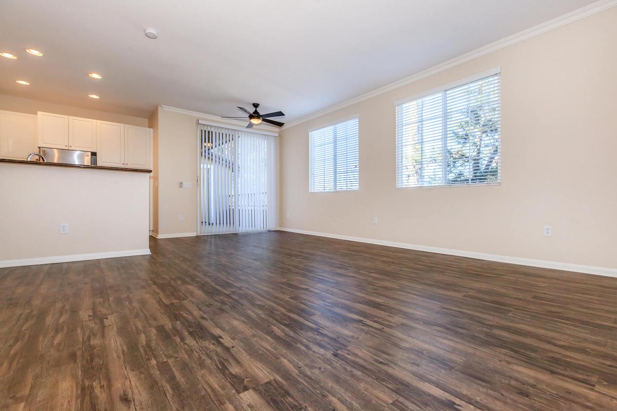 A spacious, empty living area featuring light-colored walls and wooden flooring. There are large windows with white blinds, allowing natural light to illuminate the room. A ceiling fan hangs from the ceiling, and an open kitchen space is visible in the background. The atmosphere is bright and minimalistic.