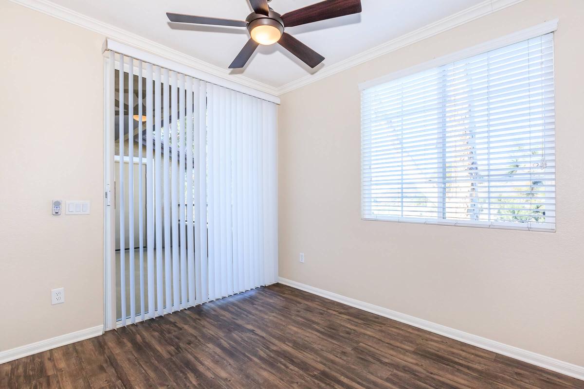 A light-colored room featuring a ceiling fan, vertical blinds, and a large window with horizontal blinds. The floor is a dark wood laminate. The space is well-lit and has a clean, minimalistic appearance, making it suitable for various purposes.