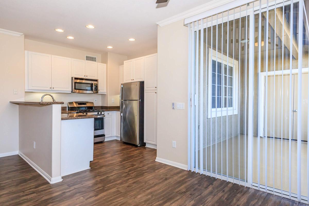 Modern kitchen with stainless steel appliances, including an oven and refrigerator, and white cabinetry. A sliding glass door leads to a sunlit room with large windows. The flooring is a warm wood tone, and the walls are painted a soft, neutral color. The space is bright and inviting.