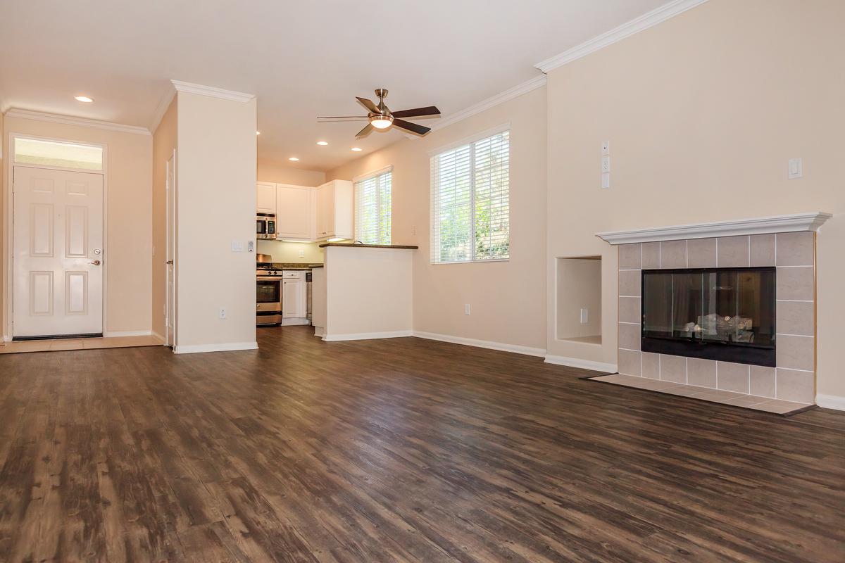 A spacious living room with a modern design, featuring hardwood flooring, large windows allowing natural light, and a ceiling fan. A kitchenette is visible in the background, along with a front door. A fireplace is located on the right side, adding warmth to the space. The walls are a light beige color.