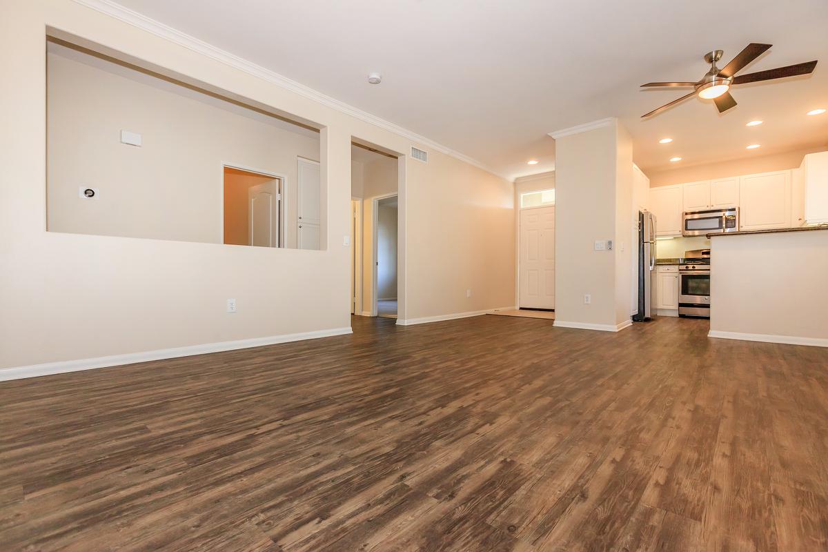 Spacious interior of a modern apartment featuring an open floor plan with hardwood-style flooring, a ceiling fan, and a kitchen area with white cabinets and stainless steel appliances. There are walls painted in neutral tones, and natural light fills the space, creating a warm, inviting atmosphere.