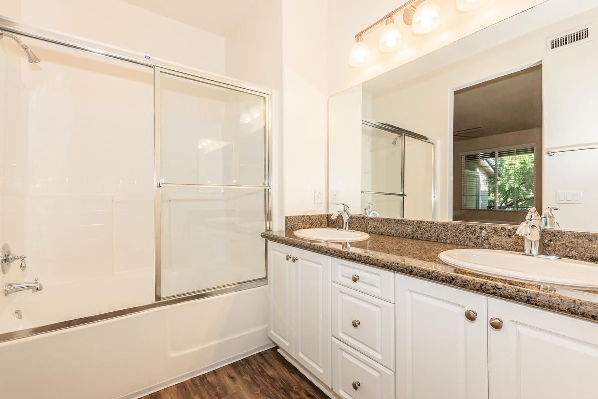 A modern bathroom featuring a double sink vanity with granite countertop, white cabinetry, a glass shower enclosure, and a bathtub. The space is well-lit with overhead lighting and a large mirror, complemented by a window allowing natural light.