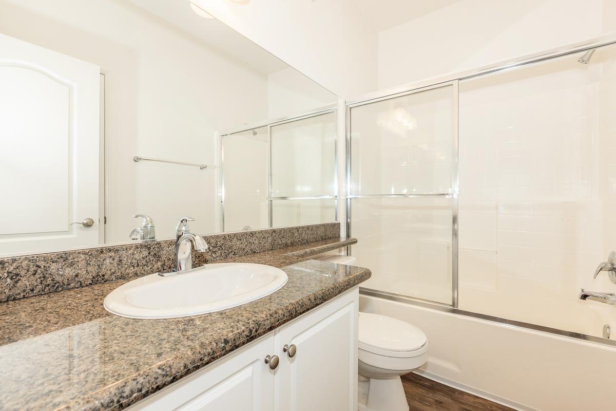 A clean, modern bathroom featuring a granite countertop sink with a mirror above, a glass-enclosed shower, a bathtub, and a white toilet. The walls are painted a light color, and the floor has a wooden appearance, creating a spacious and bright atmosphere.