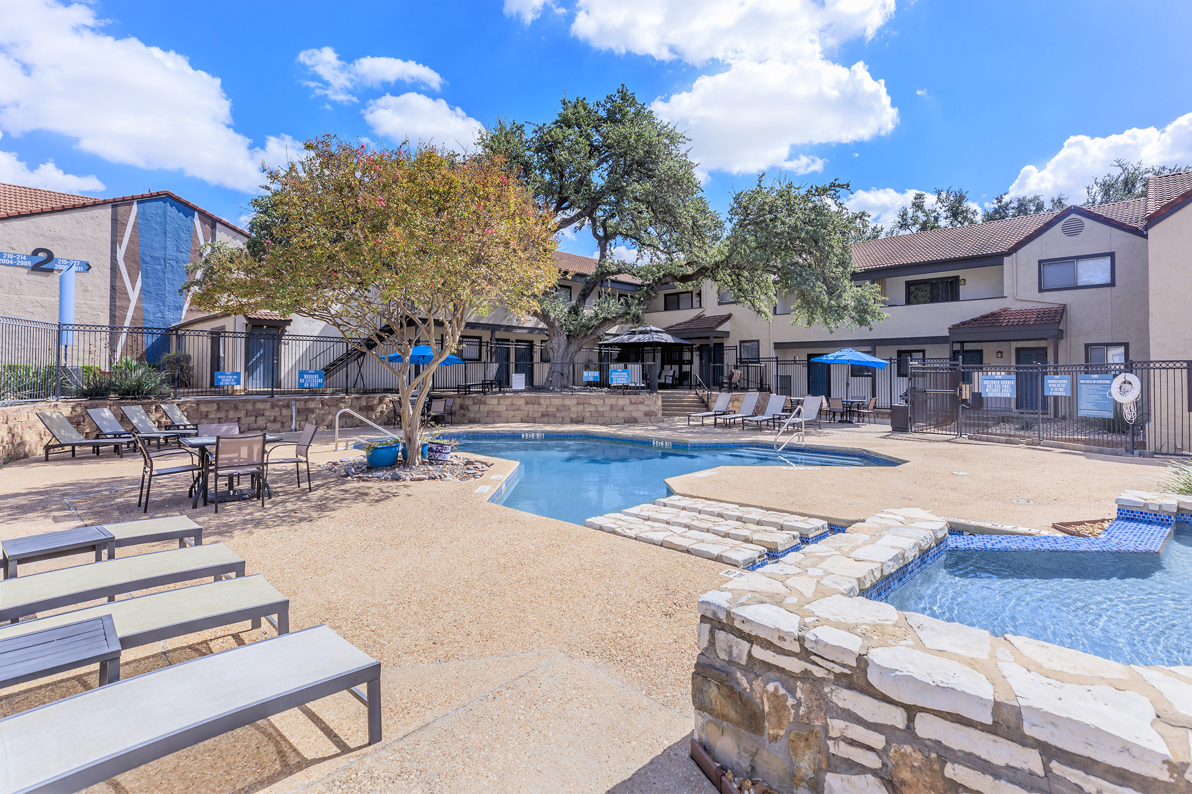 A sunny outdoor pool area featuring a curved swimming pool surrounded by lounge chairs. In the background, there are trees and apartment buildings. A hot tub is situated near the pool, creating a relaxing atmosphere. Clear blue skies and fluffy clouds enhance the cheerful setting.