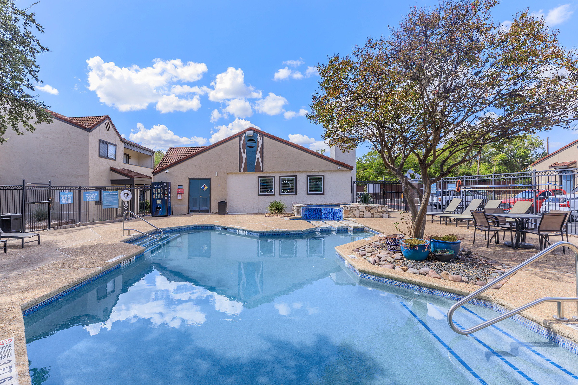 A clear blue swimming pool surrounded by lounge chairs and tables. In the background, there is a building with a mural and a fence. Lush greenery and a tree add to the landscape. The sky is bright with fluffy white clouds.