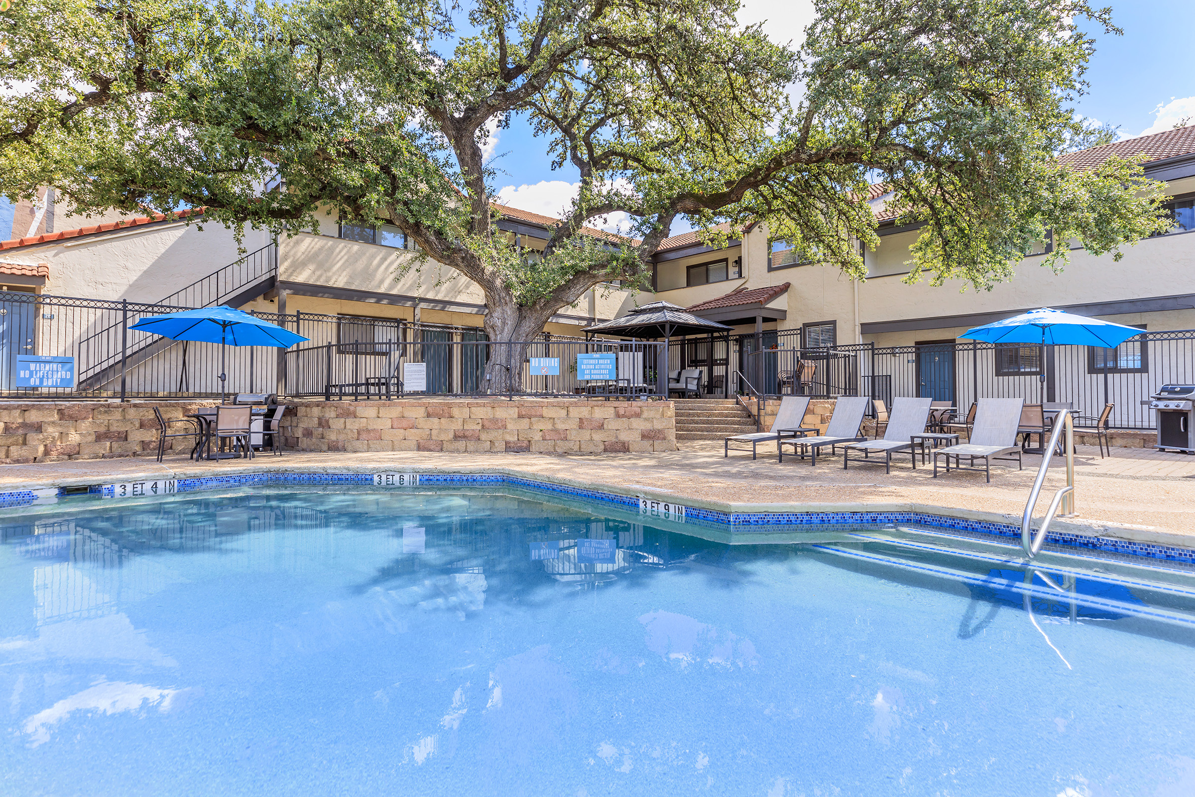 A tranquil pool area featuring clear blue water surrounded by lounge chairs and umbrellas. In the background, a building with a patio is partially shaded by a large tree. The setting is inviting and ideal for relaxation, with a sunny sky above.
