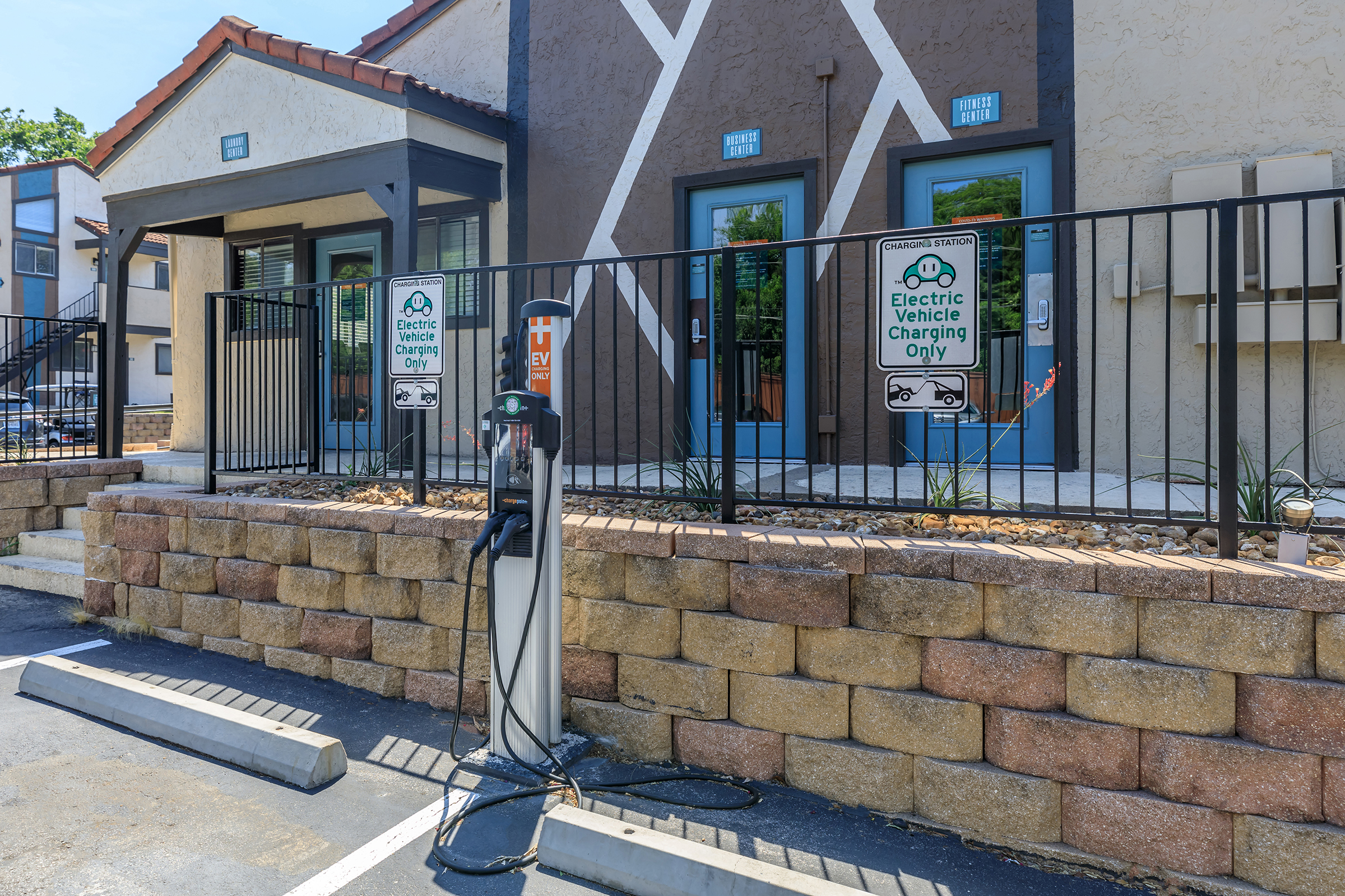 A view of a parking area featuring a charging station for electric vehicles. The station is flanked by two entrances to a building, with signs indicating "Electric Vehicle Charging Only." The surrounding area has a stone wall and decorative plants, situated under a clear blue sky.