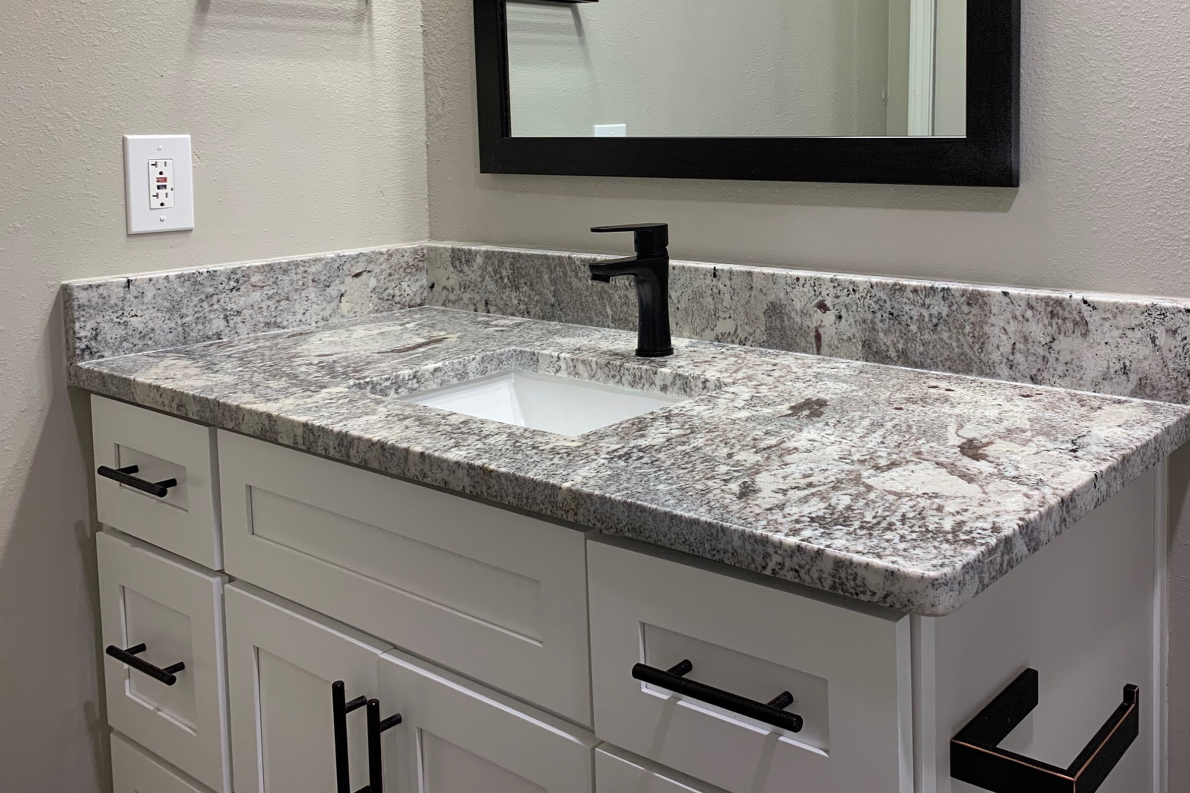 A modern bathroom vanity featuring a gray granite countertop with a white sink and a sleek black faucet. The cabinetry is white with simple black handles. A large rectangular mirror with a black frame is mounted above the sink, and the wall is painted a light gray.