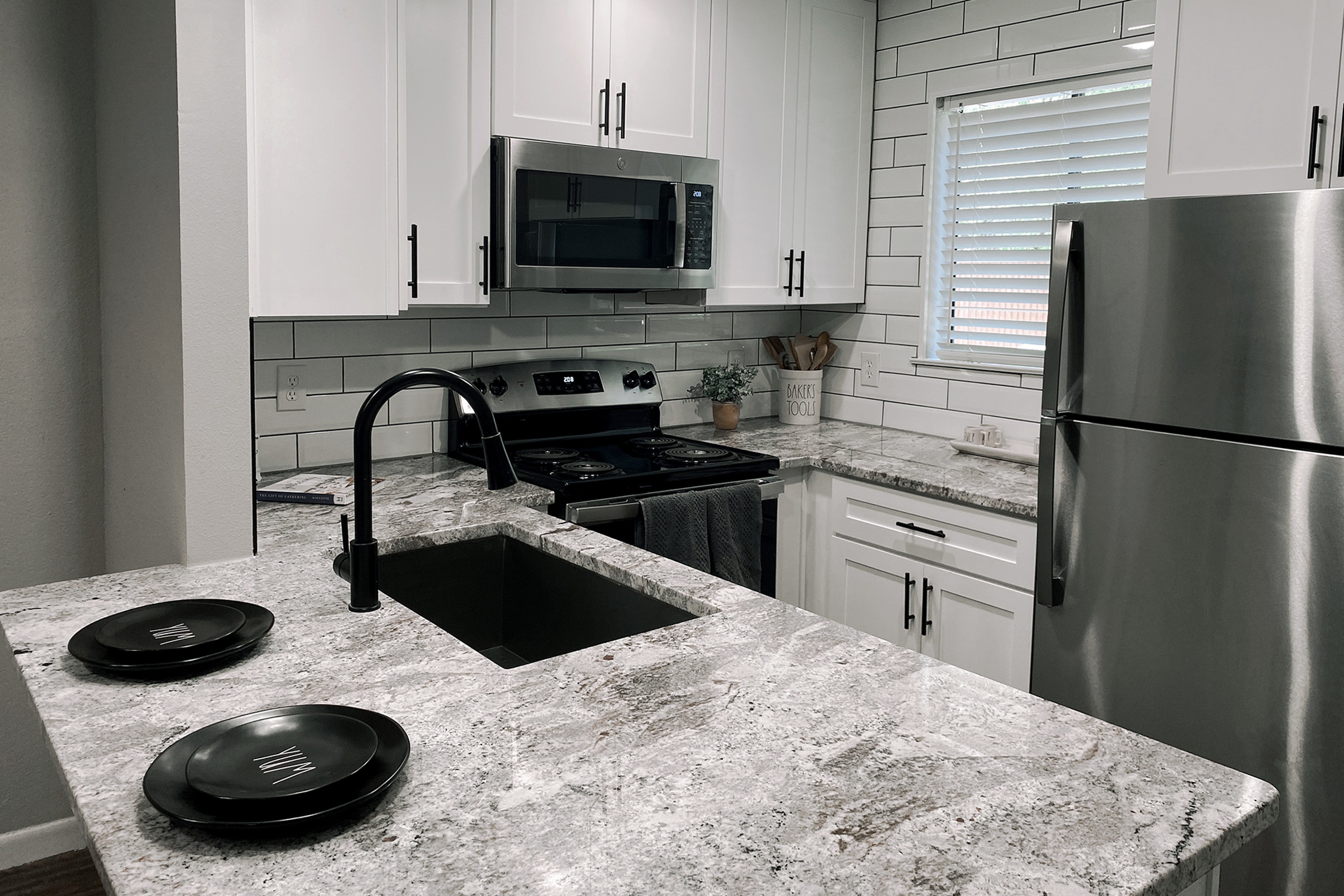 Modern kitchen featuring white cabinetry, stainless steel appliances, and a large granite countertop. A double sink with a sleek black faucet is in the foreground, alongside two black plates. The backsplash consists of white tiles, and there is a window allowing natural light in.