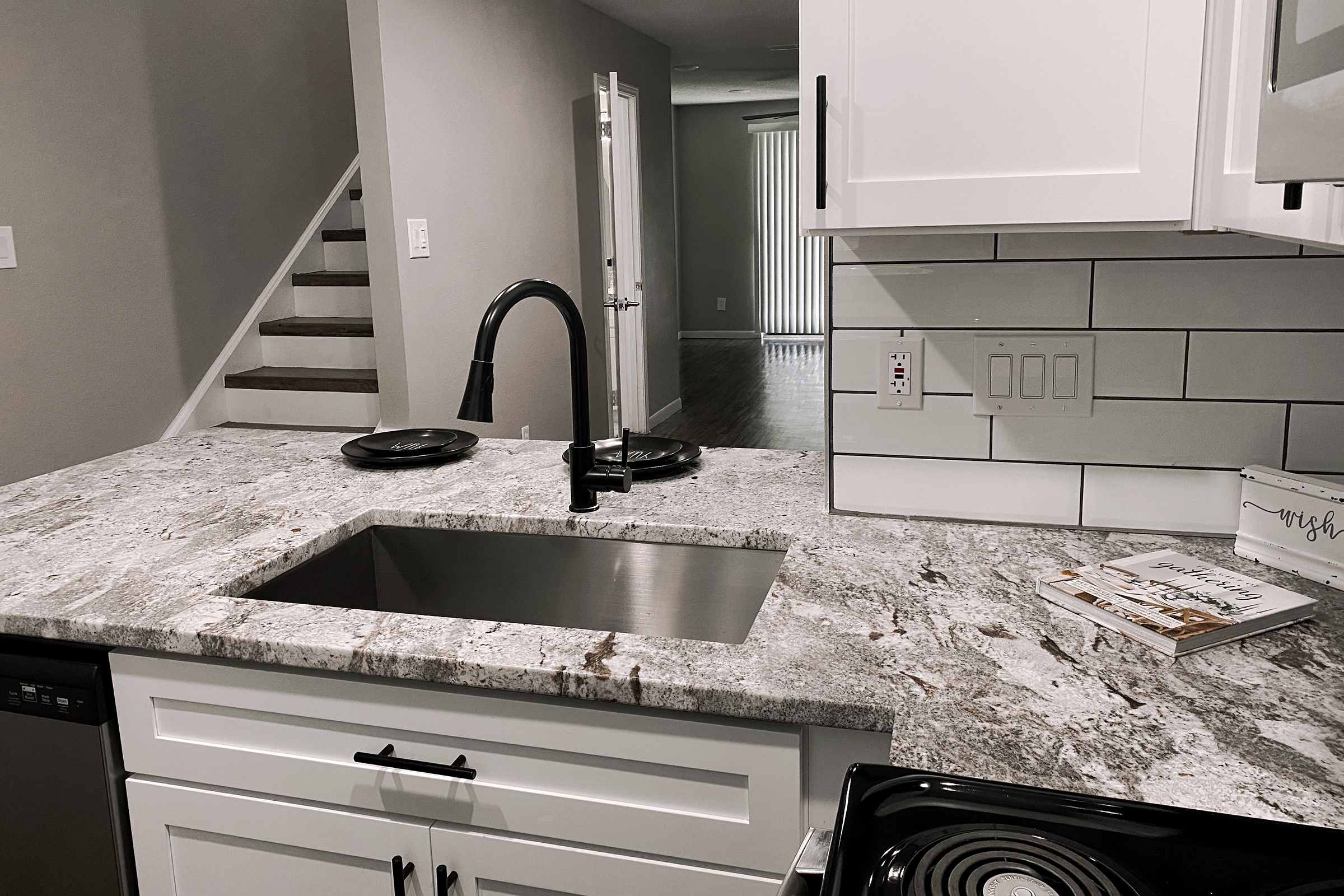 View of a modern kitchen featuring a stainless steel sink set into a gray marble countertop. White cabinetry with black handles is visible, along with a sleek faucet. In the background, a staircase leads to a light-filled living area with wooden flooring. A decorative book lies on the counter.