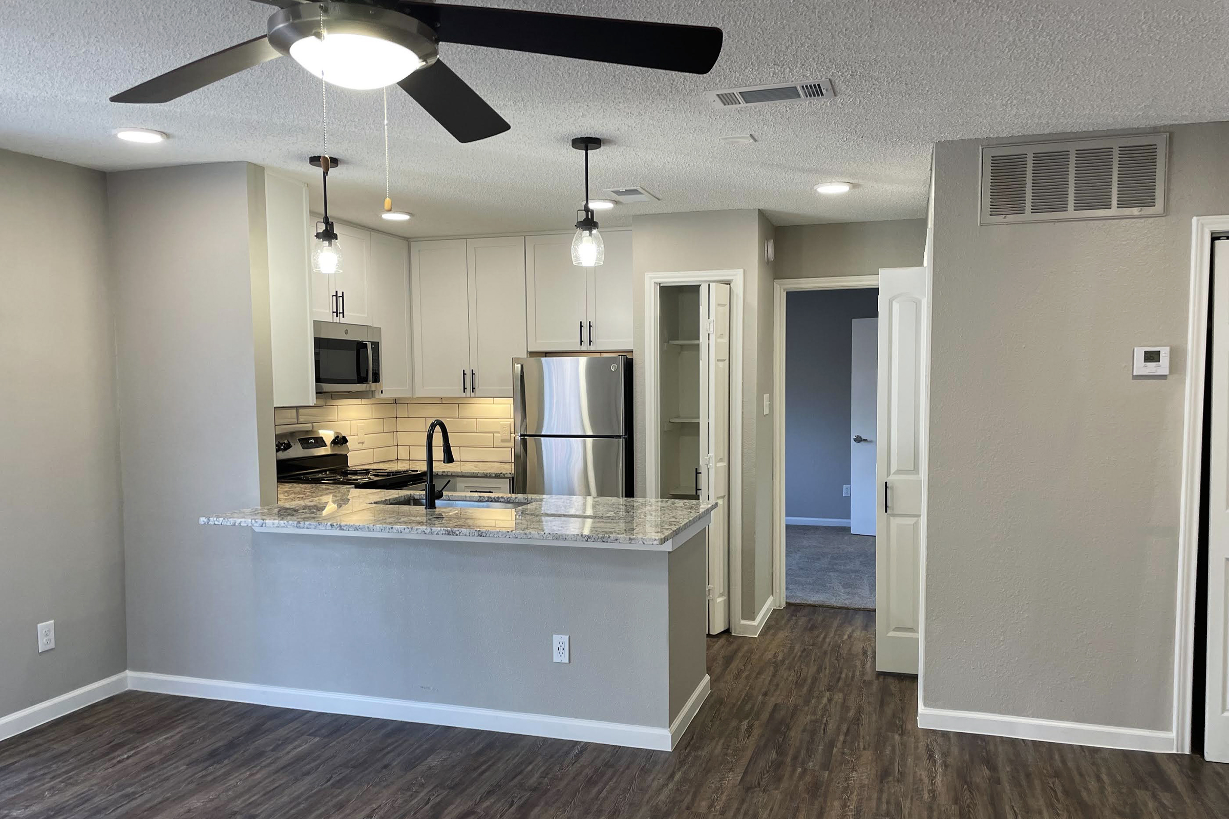 A modern kitchen and living space featuring a granite countertop with a sink, stainless steel appliances, and a ceiling fan. The area is well-lit with pendant lights. There are neutral-colored walls and a door leading to another room, showcasing a clean and updated interior design.