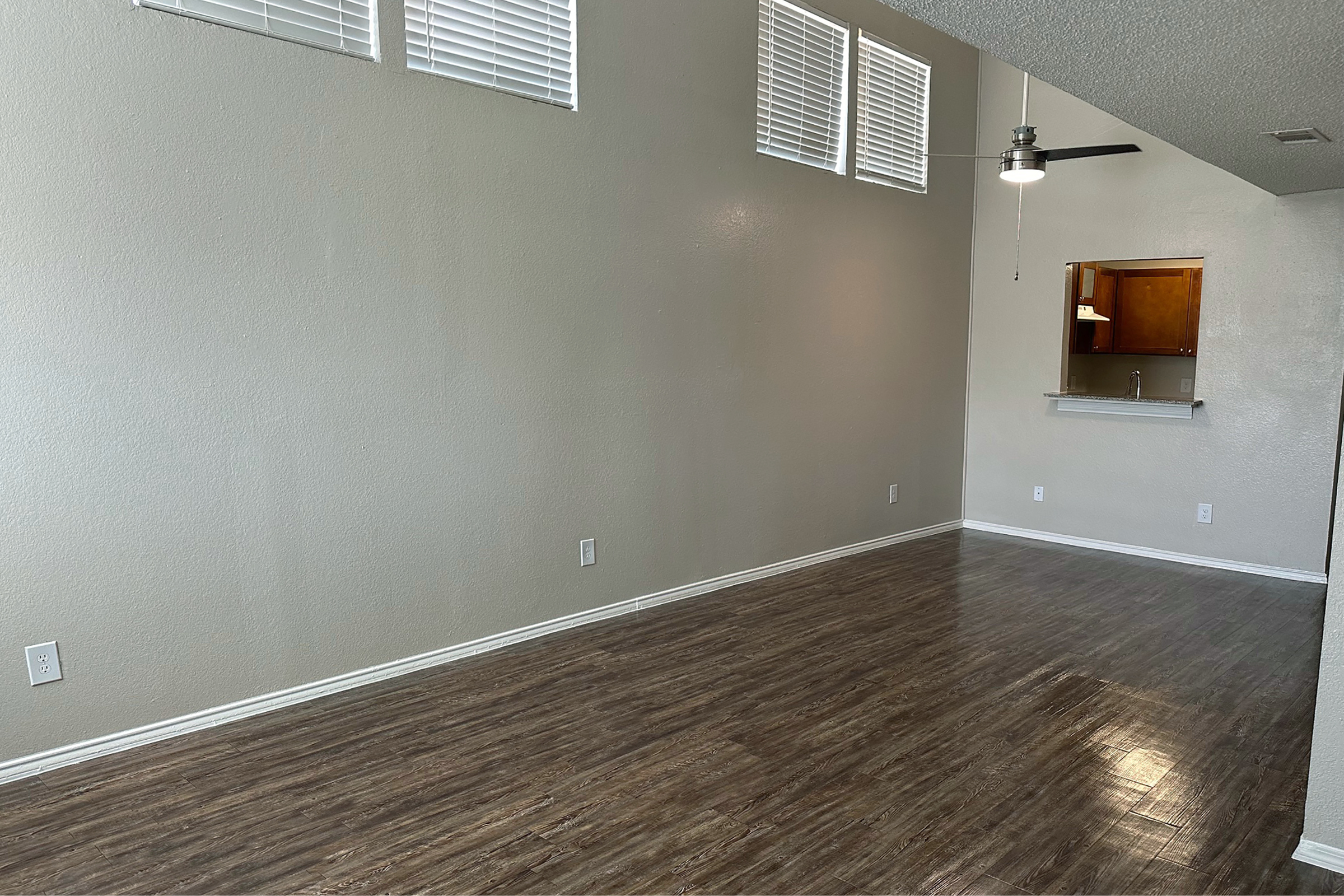 A spacious, empty living room with wooden flooring, light-colored walls, and several high windows. A ceiling fan is mounted on the ceiling. In the back, there's a small opening that leads to a kitchen area, featuring a bar counter. The room is well-lit with natural light.
