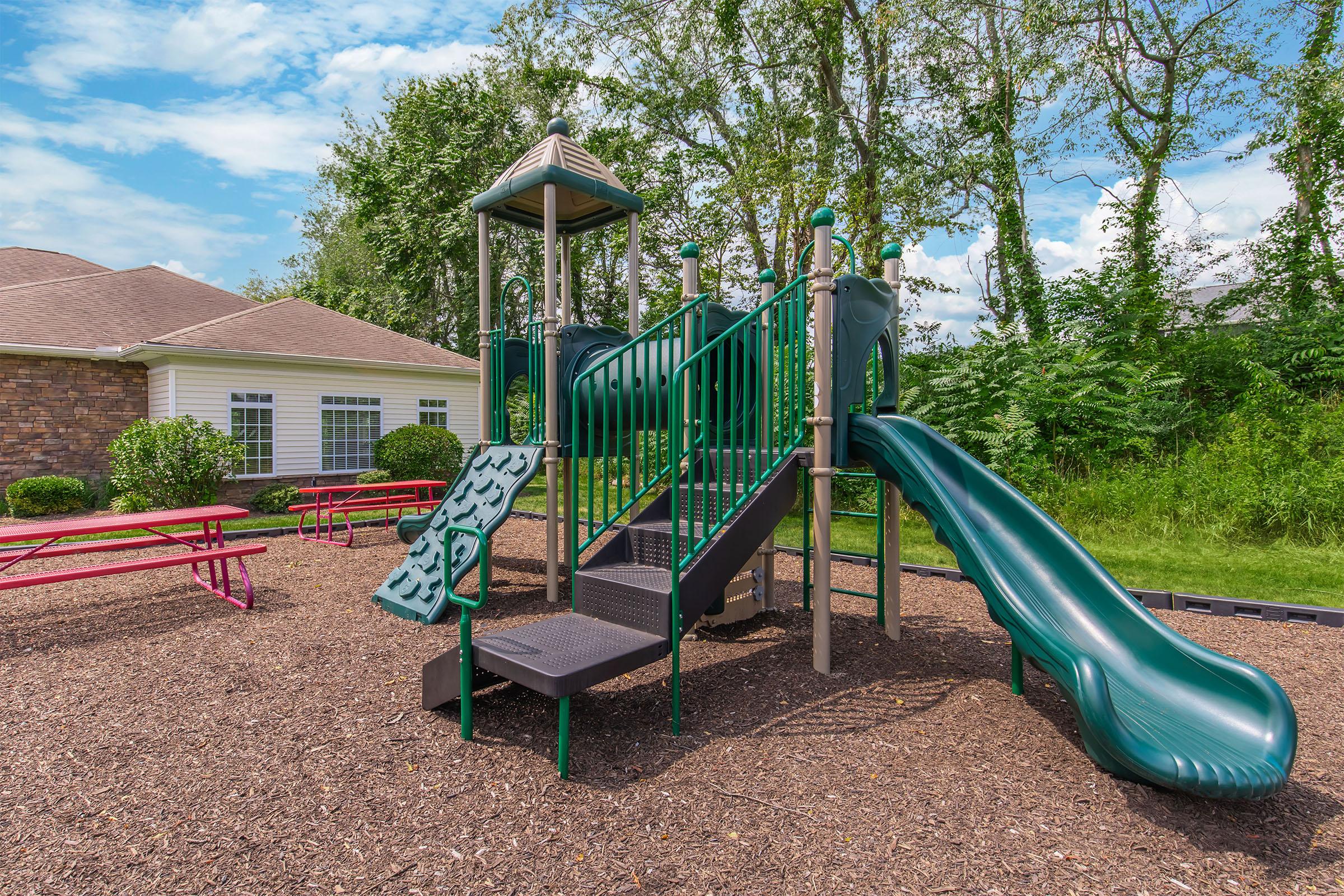 A green playground structure with slides, stairs, and climbing features, situated on a bed of mulch. In the background, there are picnic tables and a building with large windows, surrounded by trees and grassy areas under a blue sky with clouds.