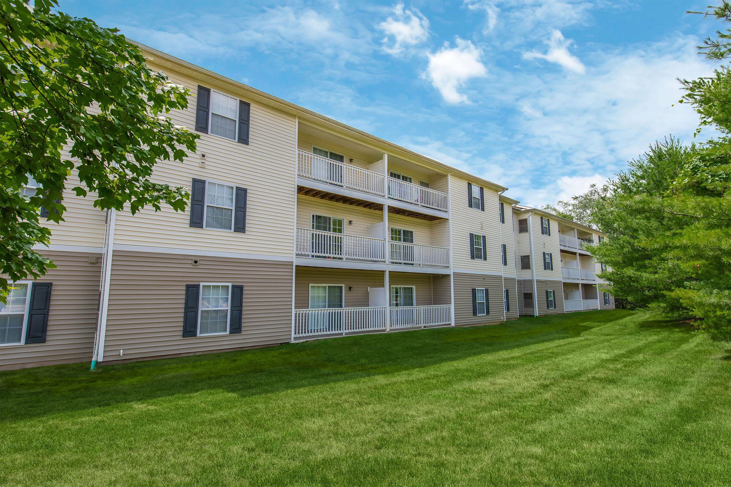 A row of beige apartment buildings featuring multiple balconies and green lawns. The sky is partly cloudy, adding to the bright and inviting atmosphere of the property. Trees line the area, enhancing the overall appeal of the residential complex.