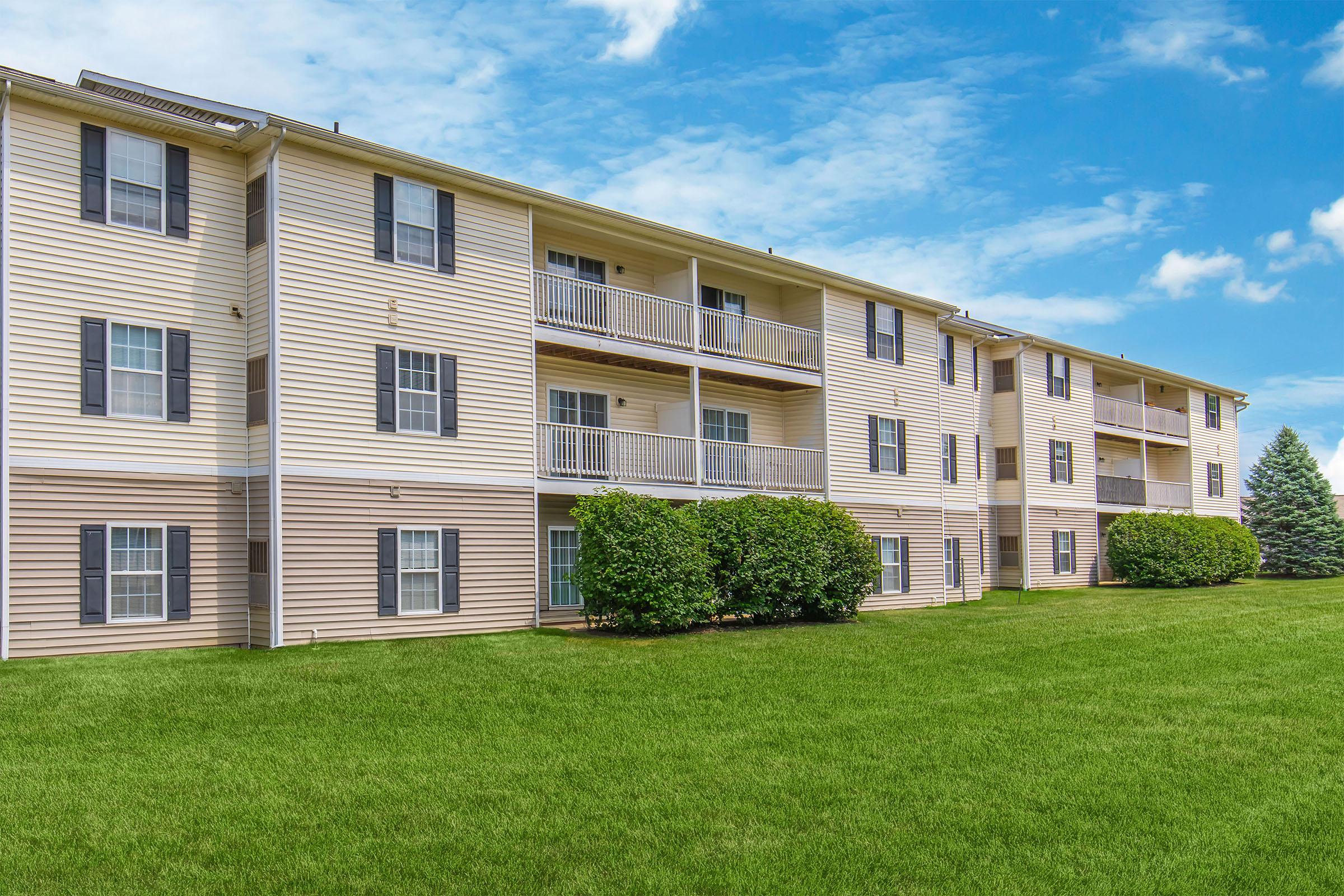 A multi-unit apartment building with beige siding, featuring multiple balconies and large windows. The building is set against a backdrop of blue skies and fluffy white clouds, surrounded by a well-maintained green lawn. Bushes are planted near the foundation, adding to the landscape.