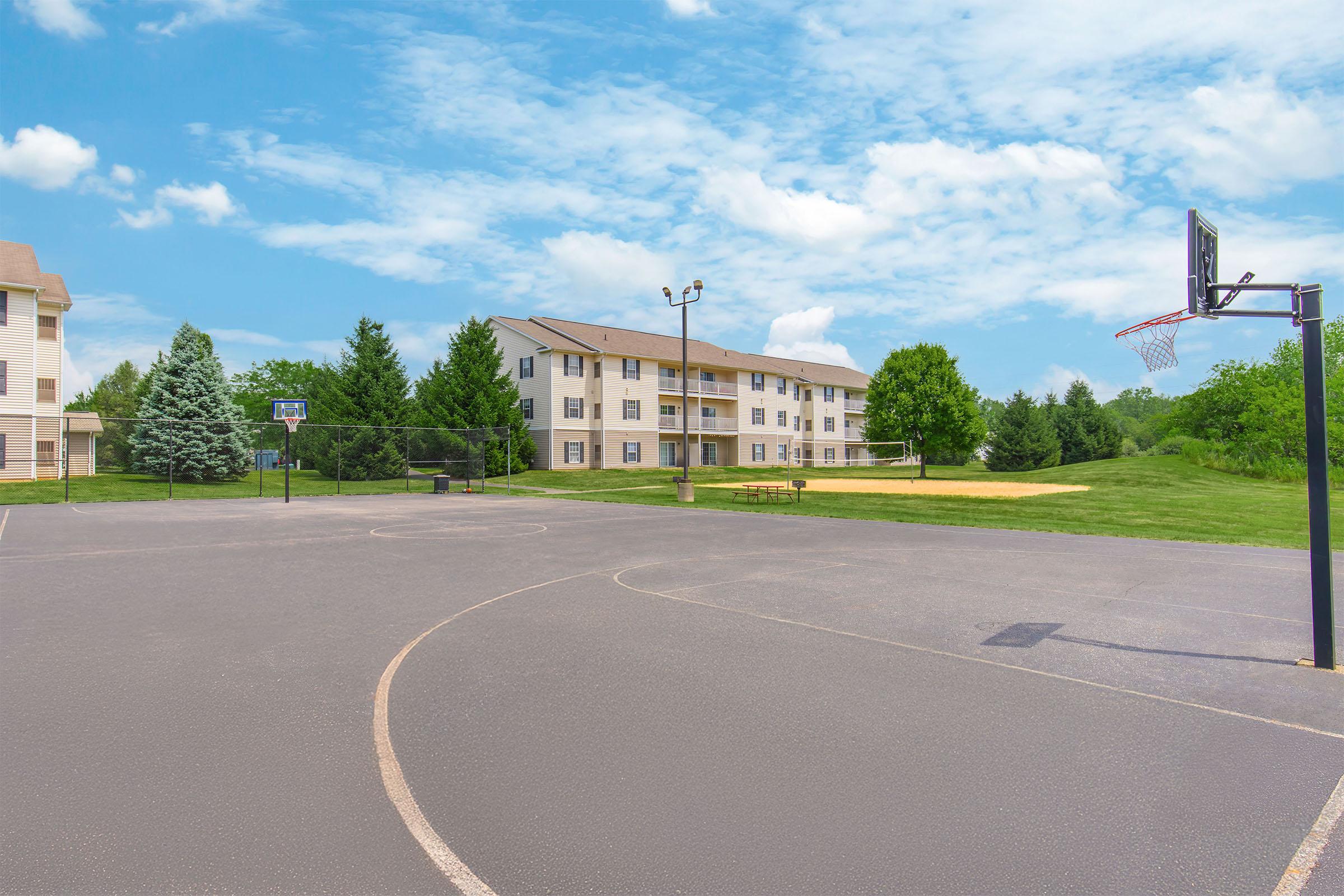 A wide view of an outdoor basketball court with a hoop, surrounded by green grass and trees. In the background, there is a multi-story residential building, under a bright blue sky with wispy clouds. The court is empty, showcasing freshly painted lines and clear surroundings.