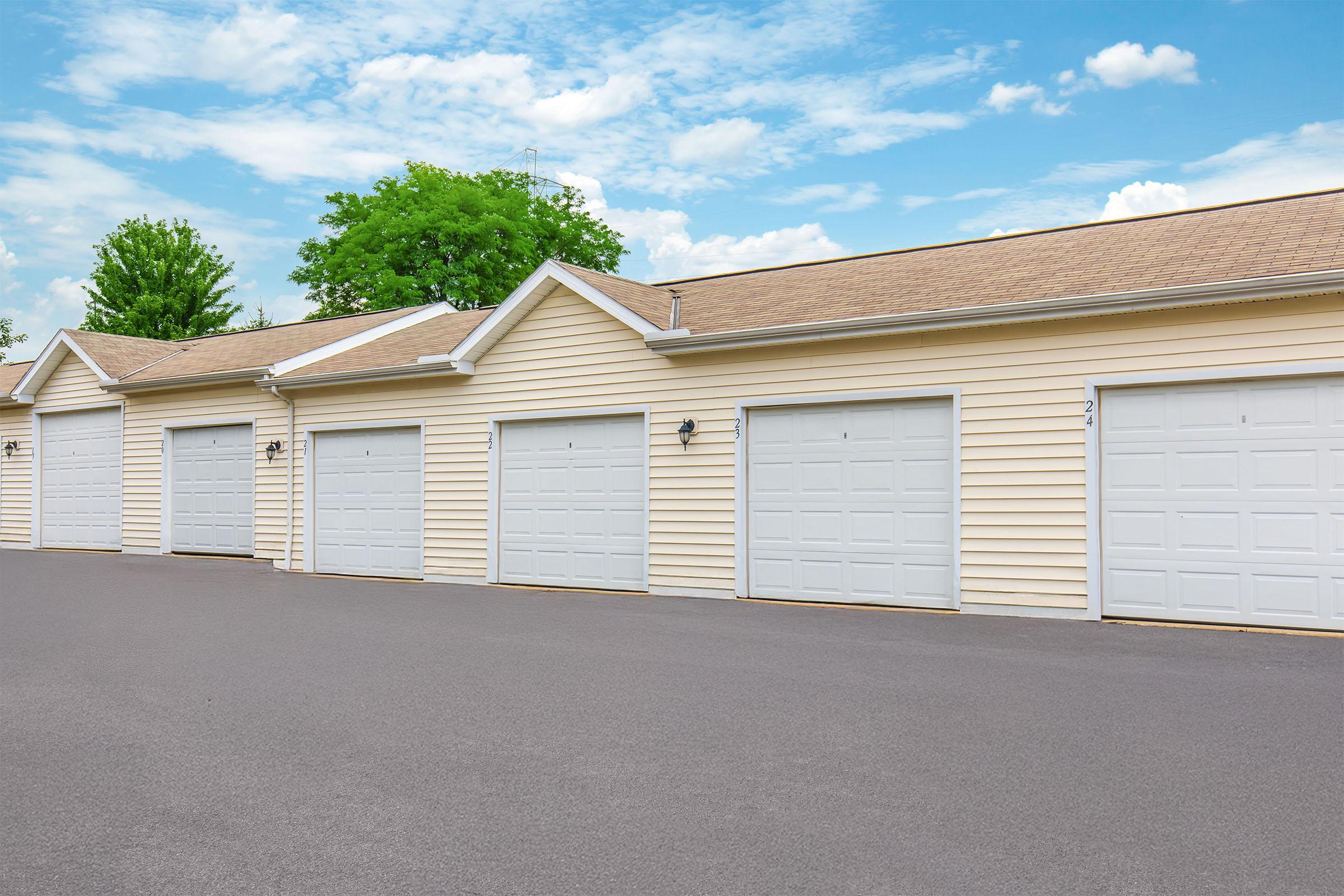A row of several closed garage doors against a light yellow building, set against a bright blue sky with scattered clouds. There are four garage doors visible in a paved area, with greenery from trees in the background. The scene suggests a suburban or residential setting.