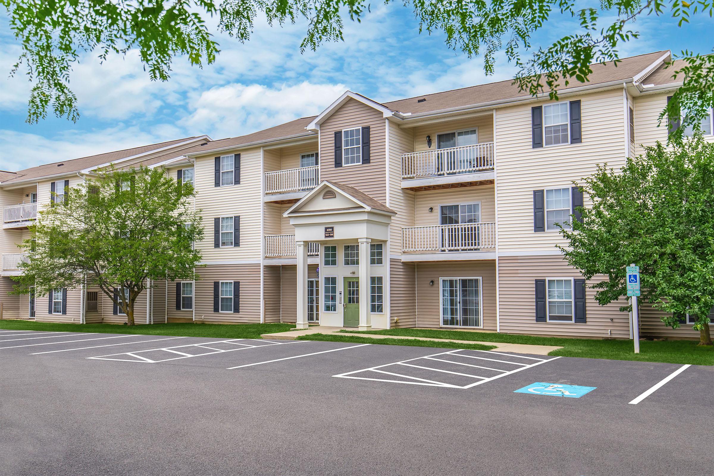 A multi-story residential building with a light beige exterior and balconies. There are several parking spaces in front, including a designated handicap spot. The surrounding area features green grass and trees, with a bright blue sky and scattered clouds above.