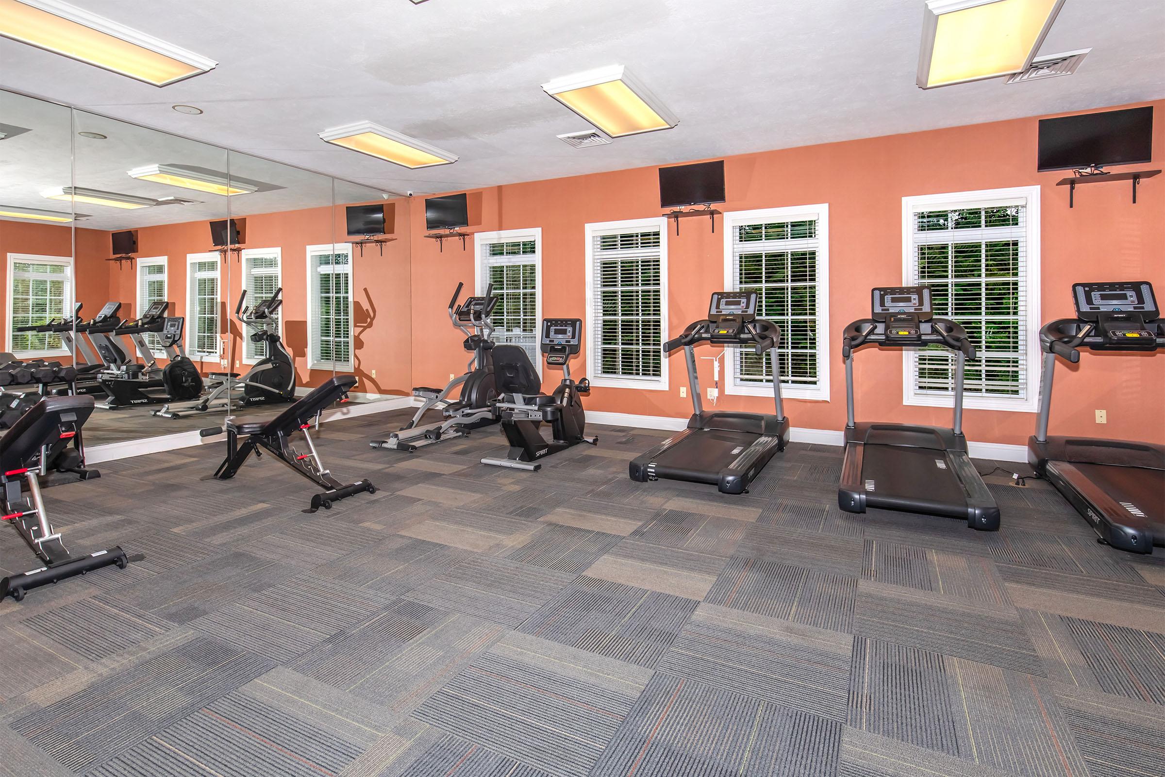A modern gym interior featuring several exercise machines, including treadmills and a stationary bike. The room has mirrored walls, ample natural light from windows, and overhead lighting. The floor is carpeted with a gray tile pattern, creating a spacious and inviting atmosphere for workouts.
